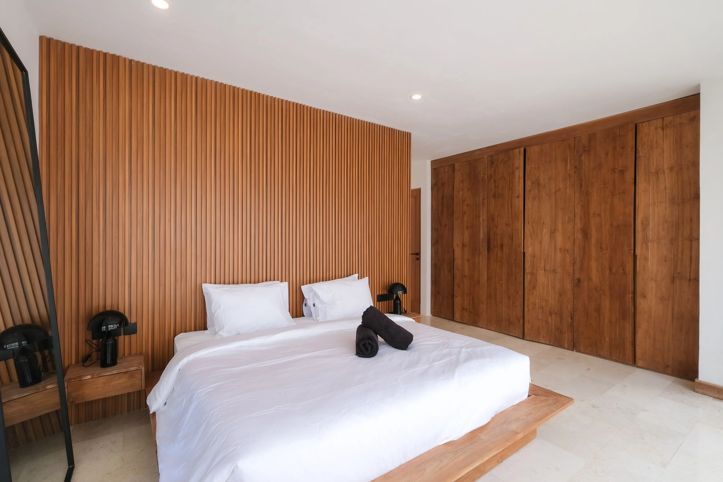 Modern bedroom with a white bed, wooden accent wall, black lamps, and large wooden closet doors.