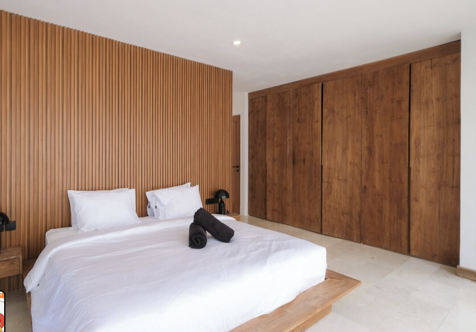 Modern bedroom with a large bed, white bedding, and two rolled black towels on top. Wooden accent wall behind the bed, wooden wardrobe to the right, and a small bedside table with a black lamp. Neutral-colored tiled floor.
