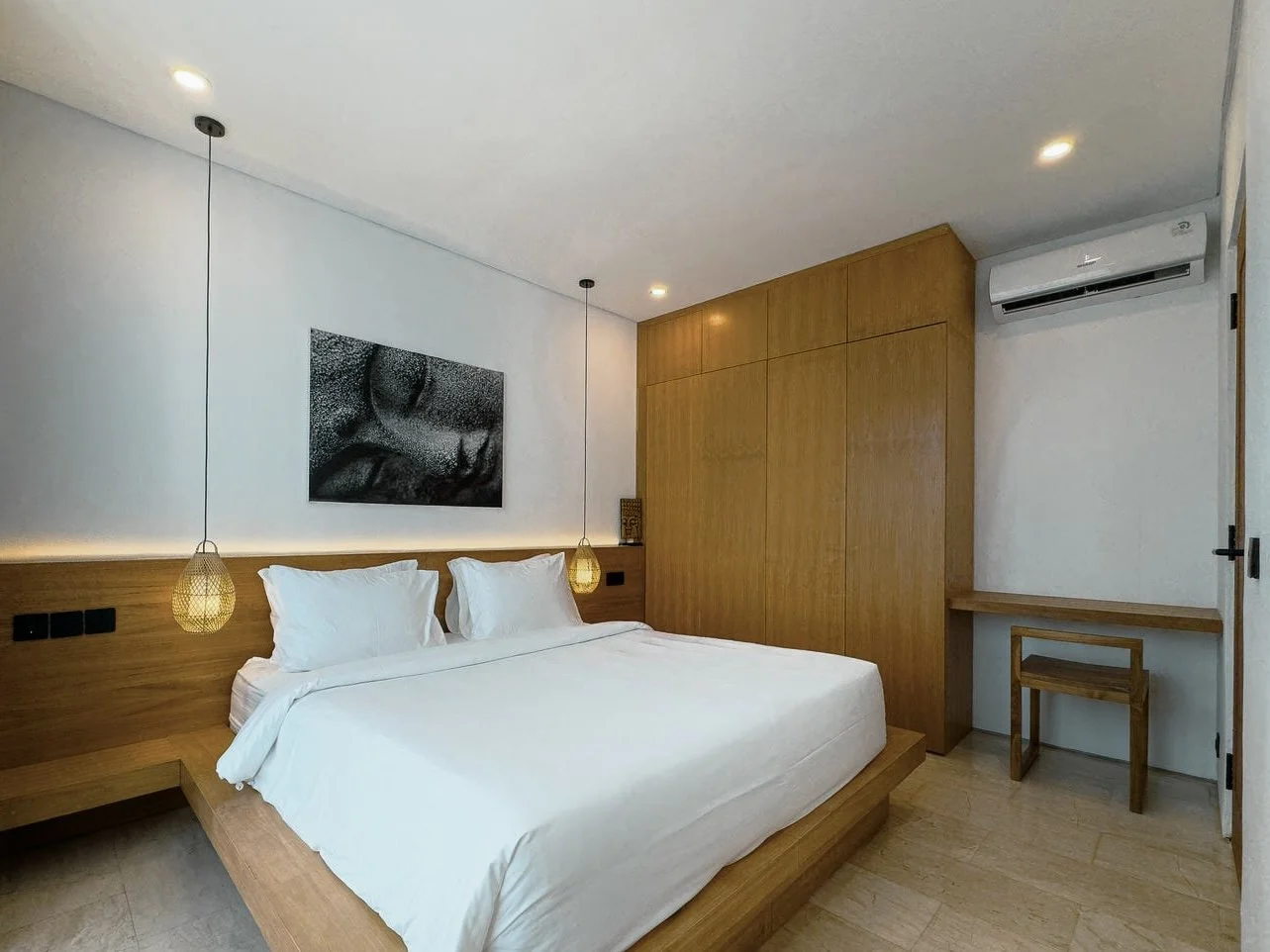 Modern hotel room with a white bed, wooden headboard, hanging pendant lights, a black and white wall art, and wooden furniture including wardrobe and a small desk.
