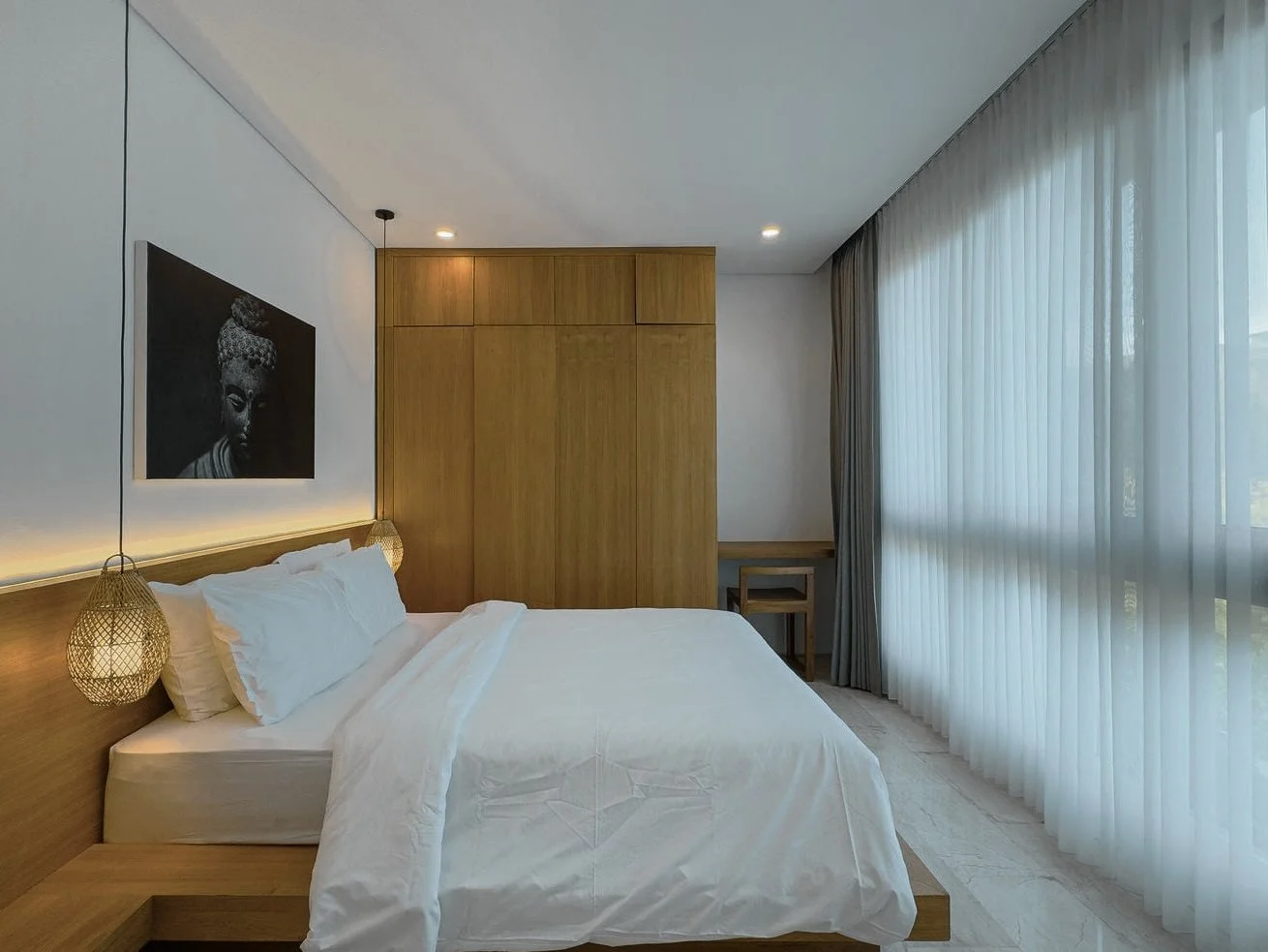 Modern bedroom with white bedding, wooden headboard, two hanging light fixtures, a black and white Buddha artwork, wooden wardrobe, a small desk with a chair, and large sheer curtains.