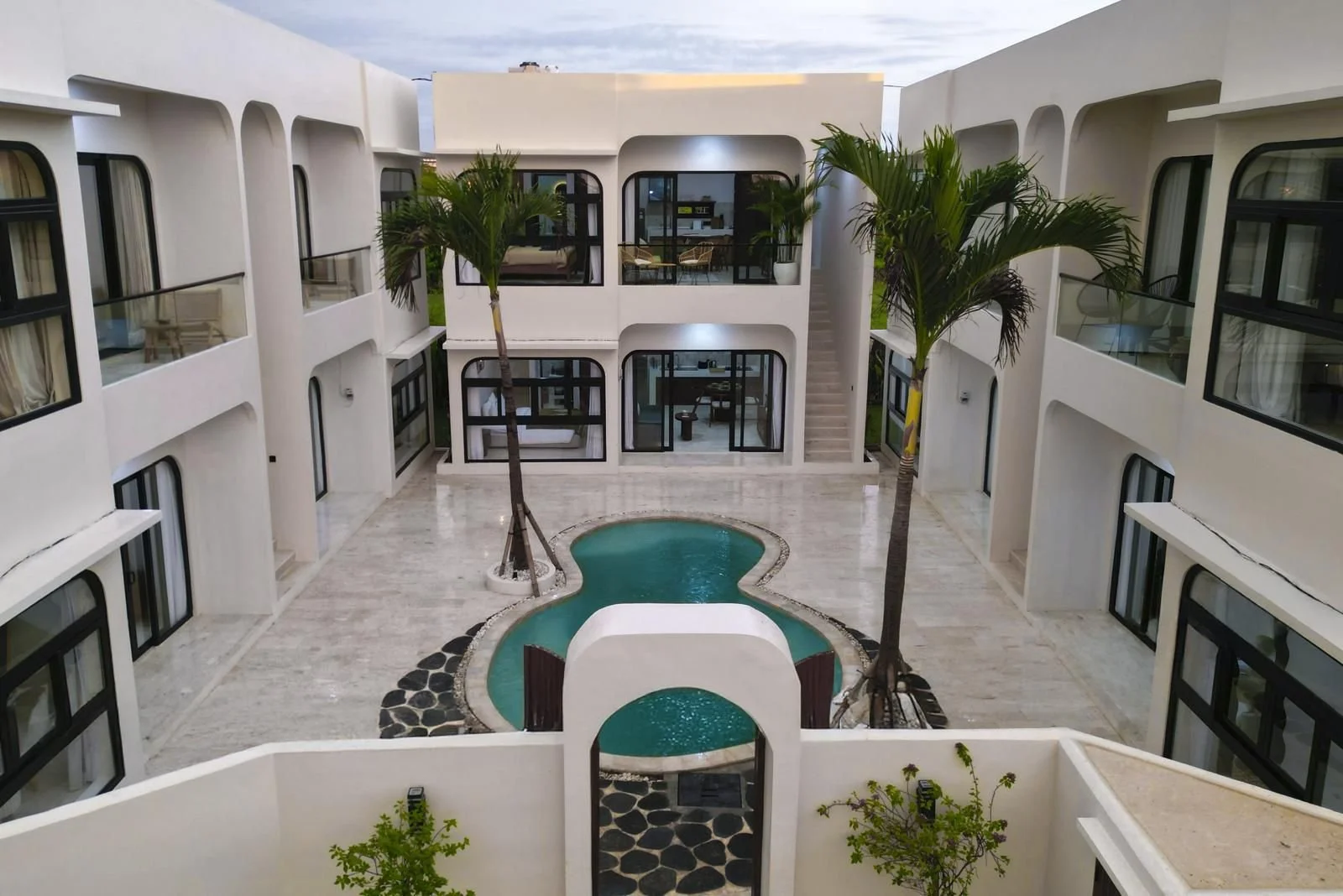 View of a modern hotel courtyard with a small pool, palm trees, and white building with large windows.