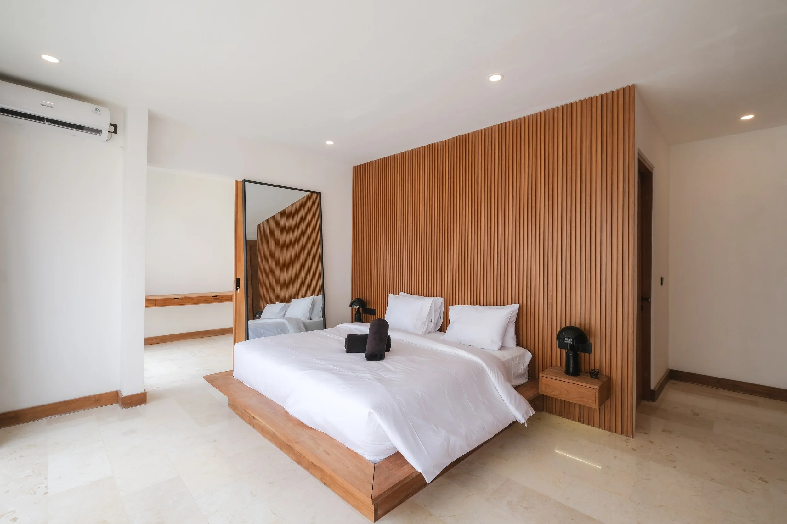 Modern bedroom with a large bed, wooden accent wall, black lamps, a full-length mirror, and minimal decor.