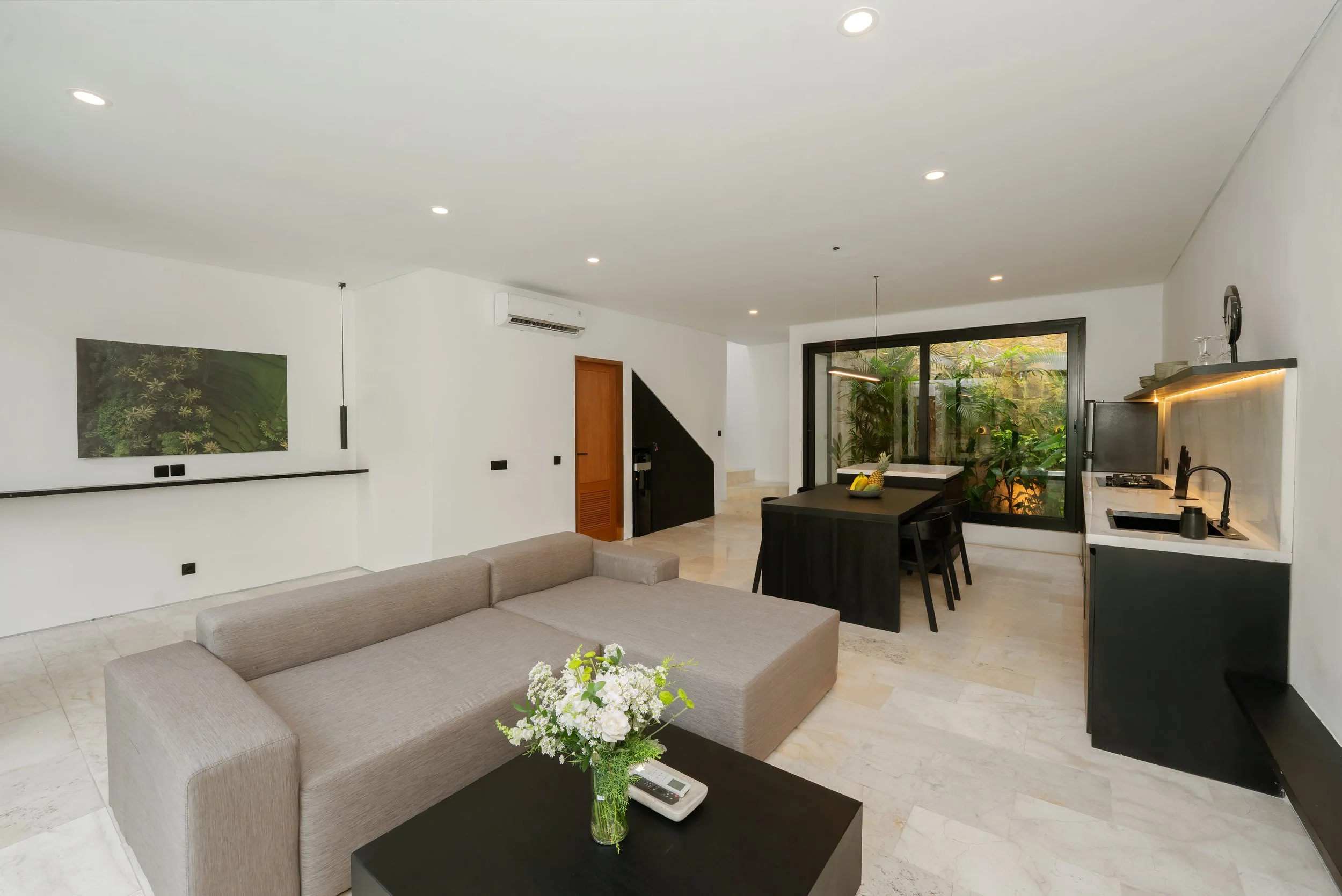 Modern living room with beige sectional sofa, black coffee table with flower vase and remote, white wall with artwork, black kitchen and dining area with window and indoor plants, staircase in background.