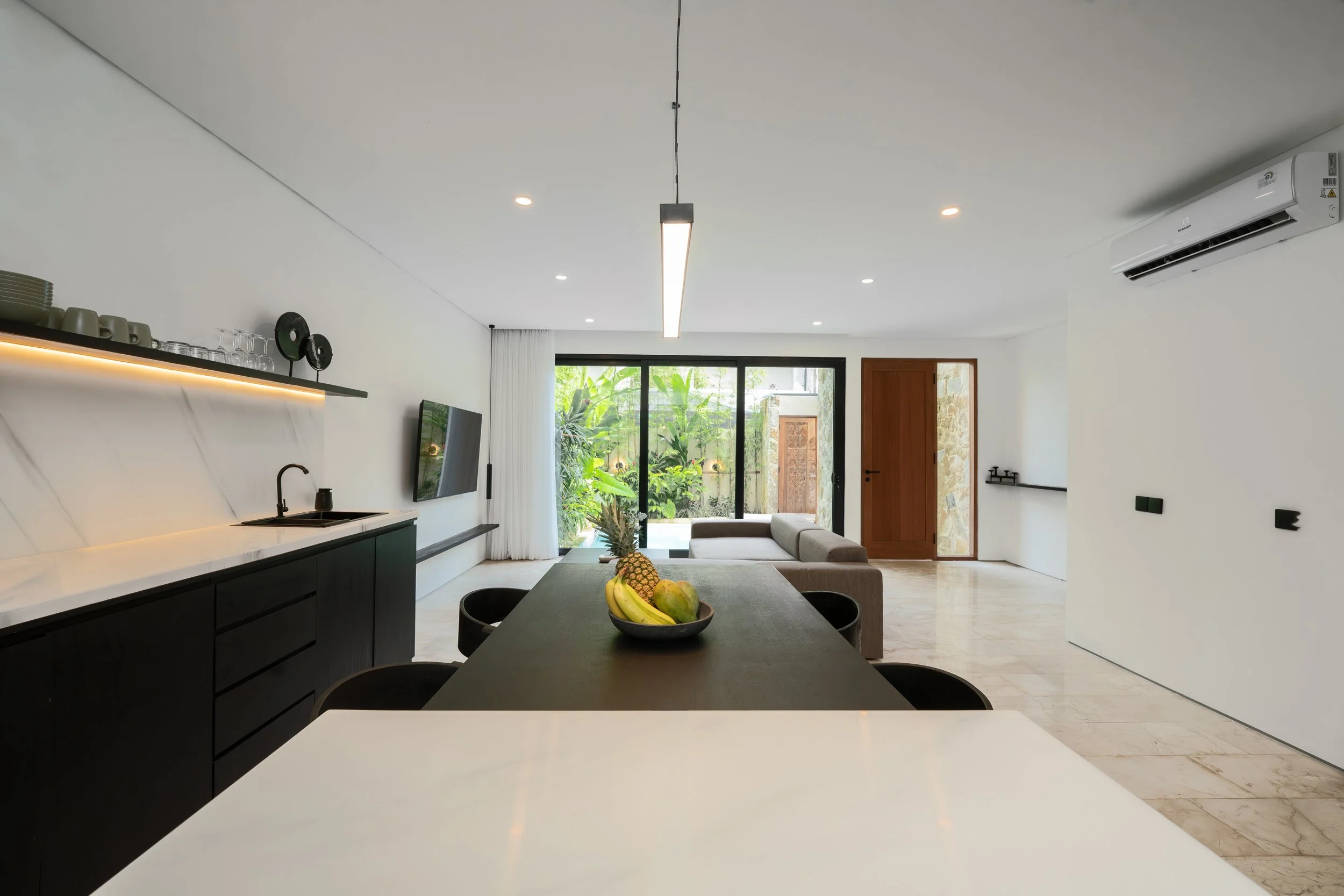 Modern open-plan living area with kitchen, dining, and sitting space, featuring a large window with greenery outside, a black dining table with a fruit bowl, and neutral decor.