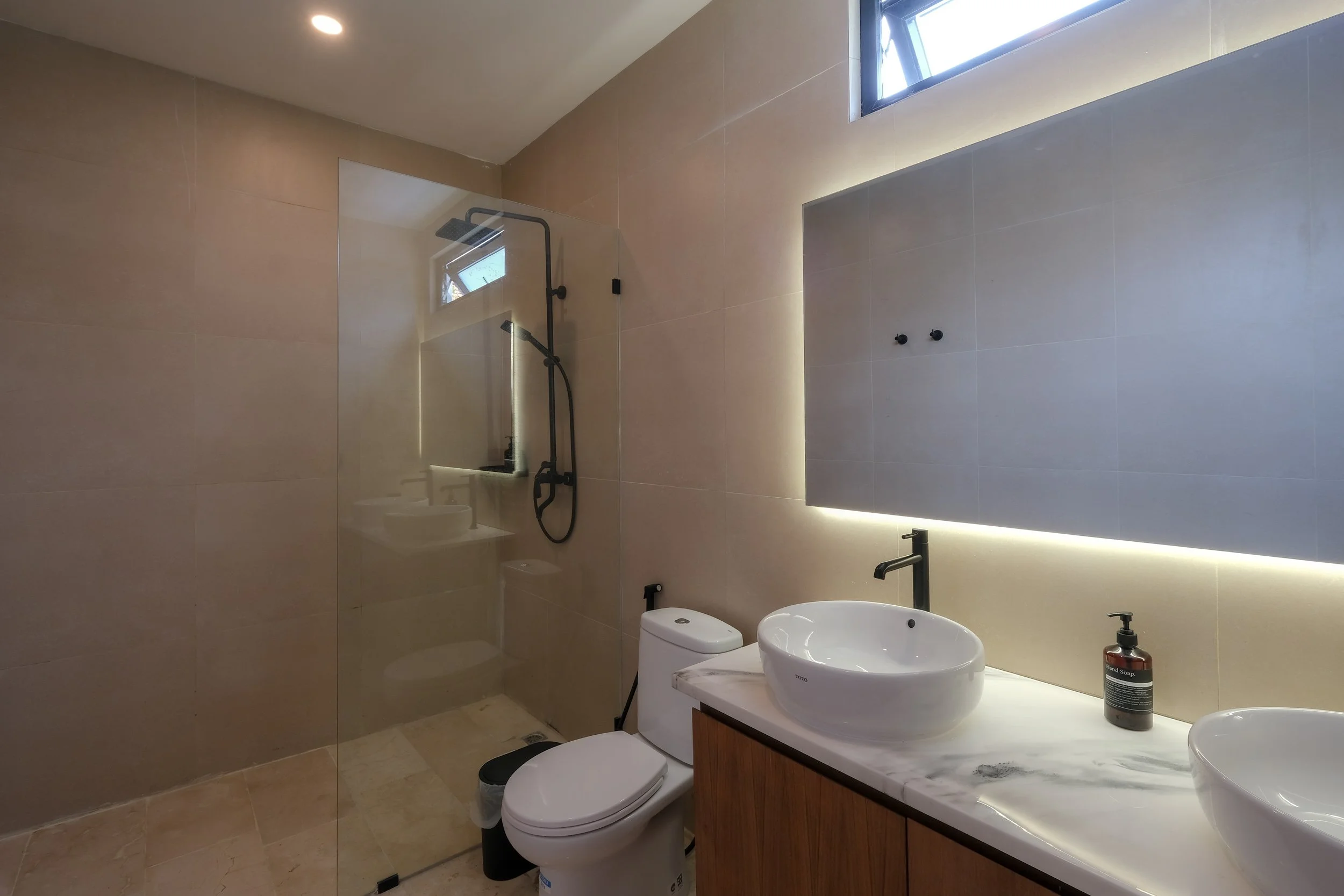 Modern bathroom with dual vessel sinks, a wall-mounted mirror with backlit lighting, a toilet, and a walk-in shower with a glass partition and black fixtures. Natural light enters through a small window.