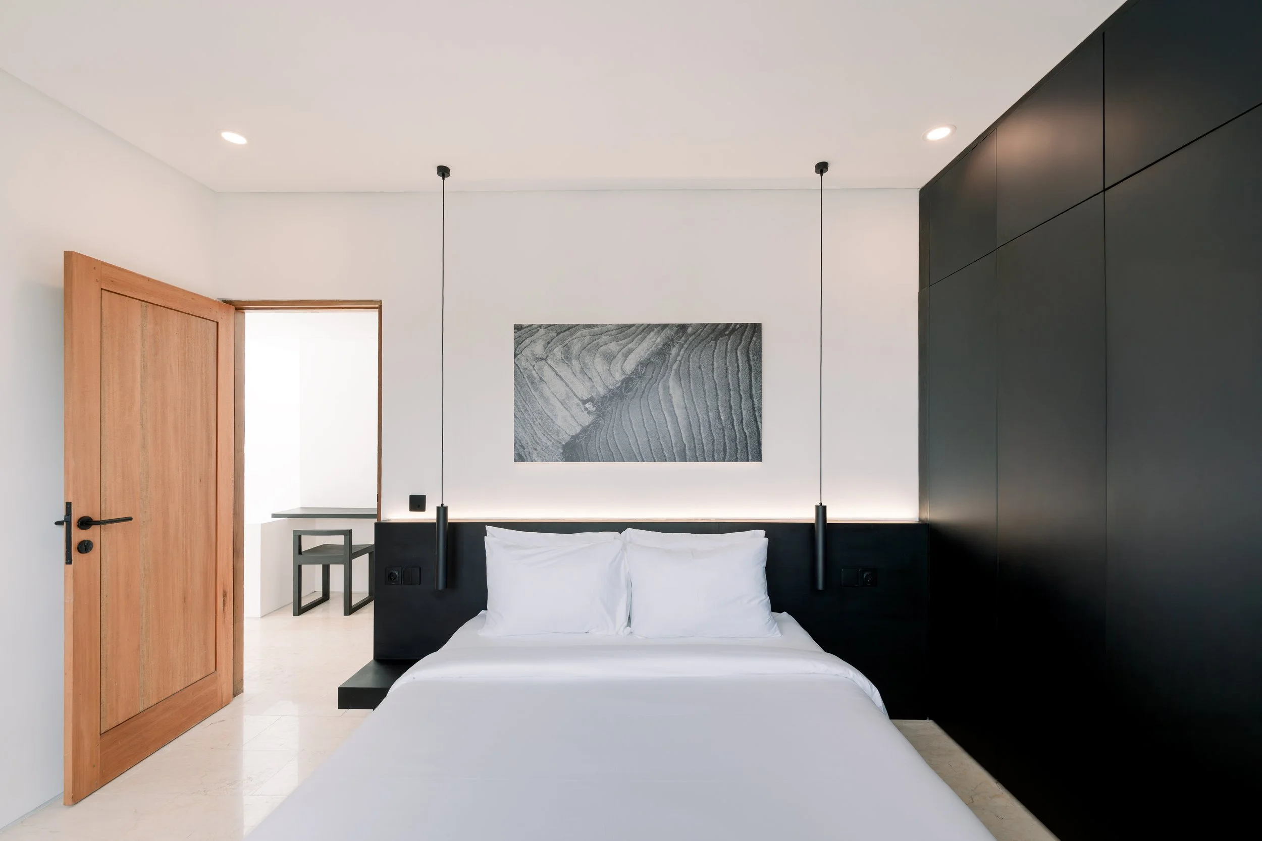 Modern bedroom with white walls, black headboard, hanging pendant lights, black wardrobe, and a wooden door.