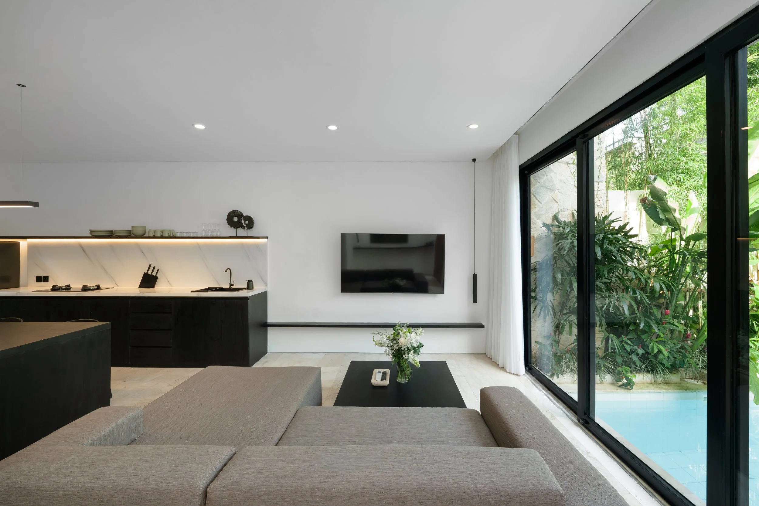 Modern living room with light-colored sofa, black coffee table with flowers and remote, wall-mounted TV, large sliding glass door showing greenery and pool outside, minimalist kitchen with black cabinets and white backsplash.