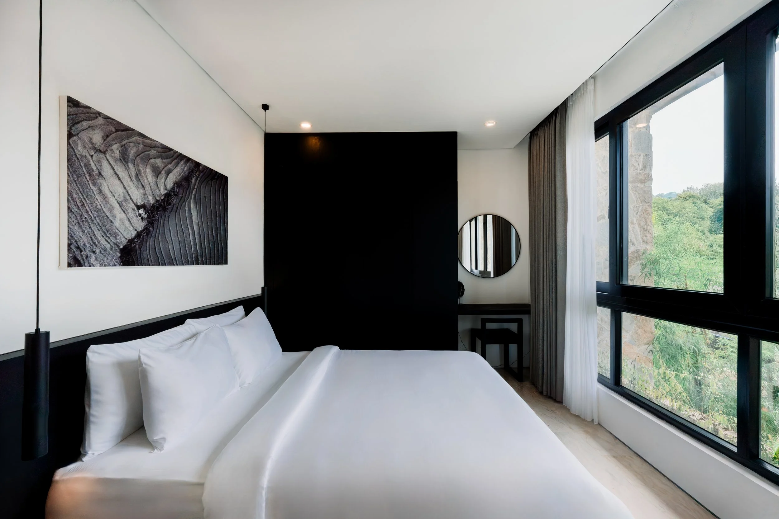 Modern hotel bedroom with white bed, black headboard, abstract artwork, black and white color scheme, large window with trees outside, circular mirror, small black desk and stool.