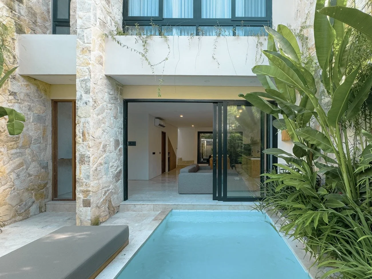 Modern stone house with a small swimming pool and sliding glass doors opening to a living room with a sofa, surrounded by green plants.