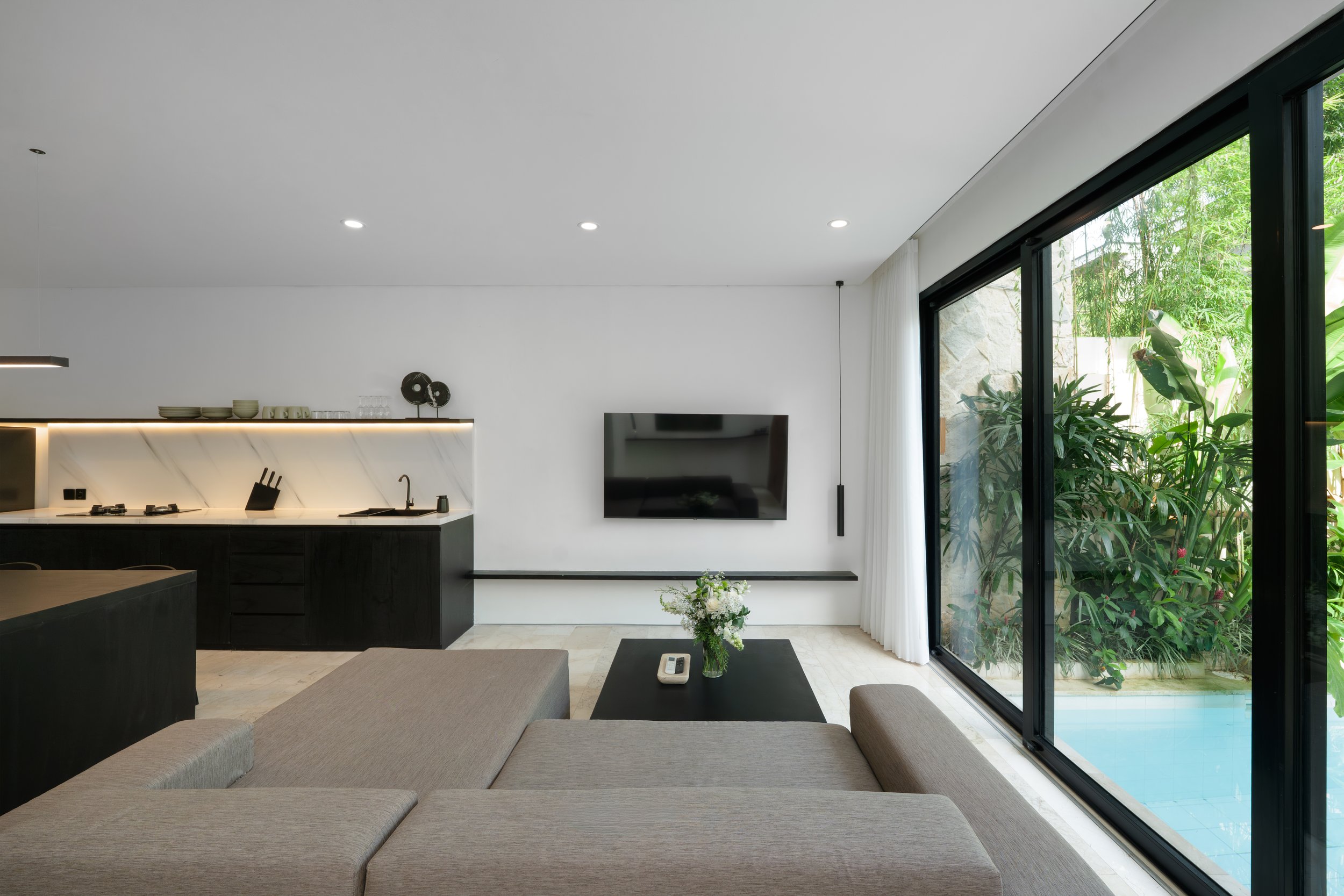 Modern living room with a beige sofa, black coffee table with a remote and flower vase, wall-mounted TV, kitchen with black cabinets and backsplash, large glass sliding door revealing outdoor greenery, and white curtains.