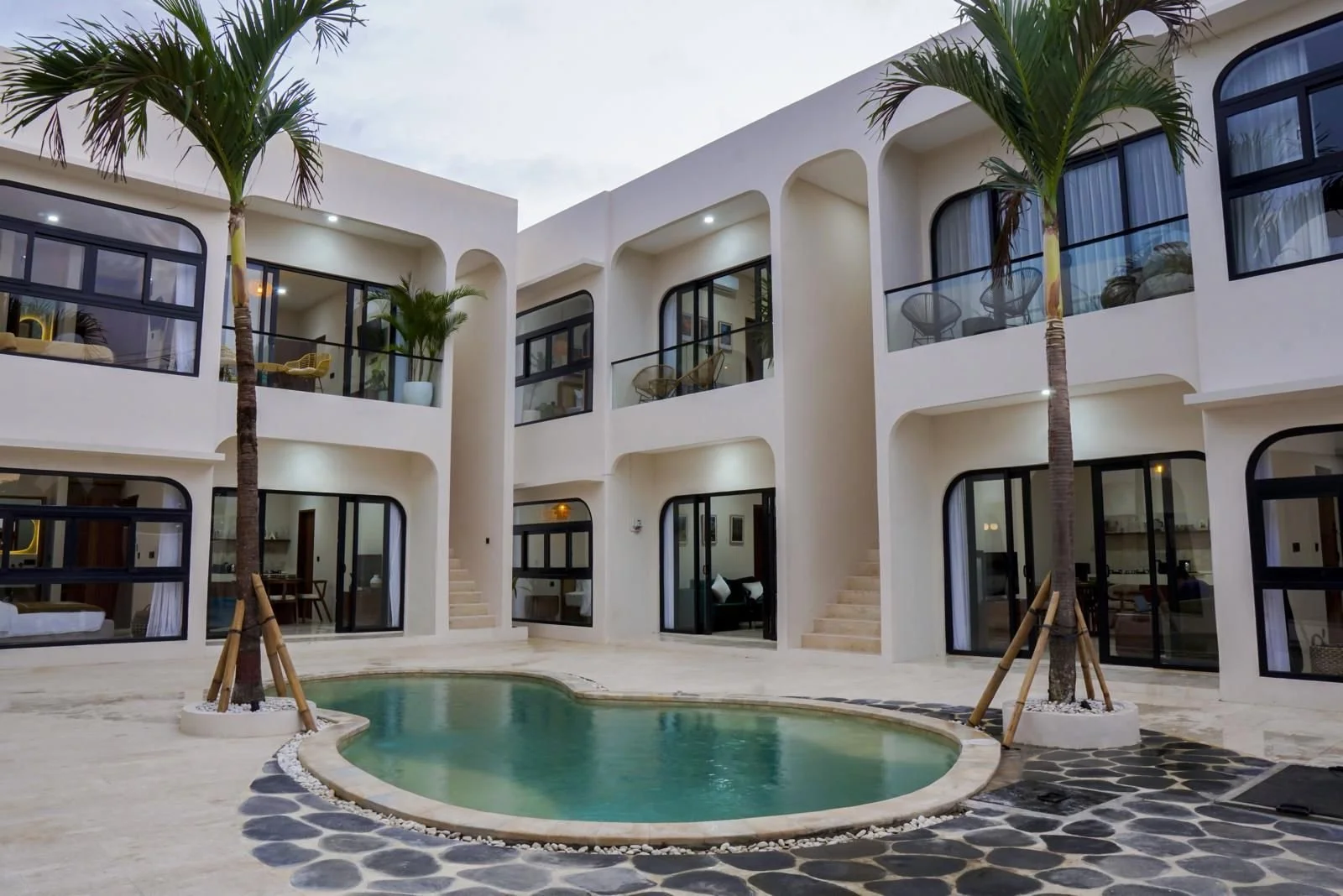 Modern white apartment building with multiple balconies, large glass doors, a small swimming pool in the courtyard, and two palm trees.