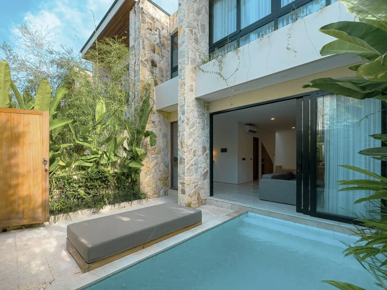 Modern house with a stone exterior, large sliding glass doors, and a small swimming pool with a cushioned lounge on the poolside. Green plants are visible around the patio area.