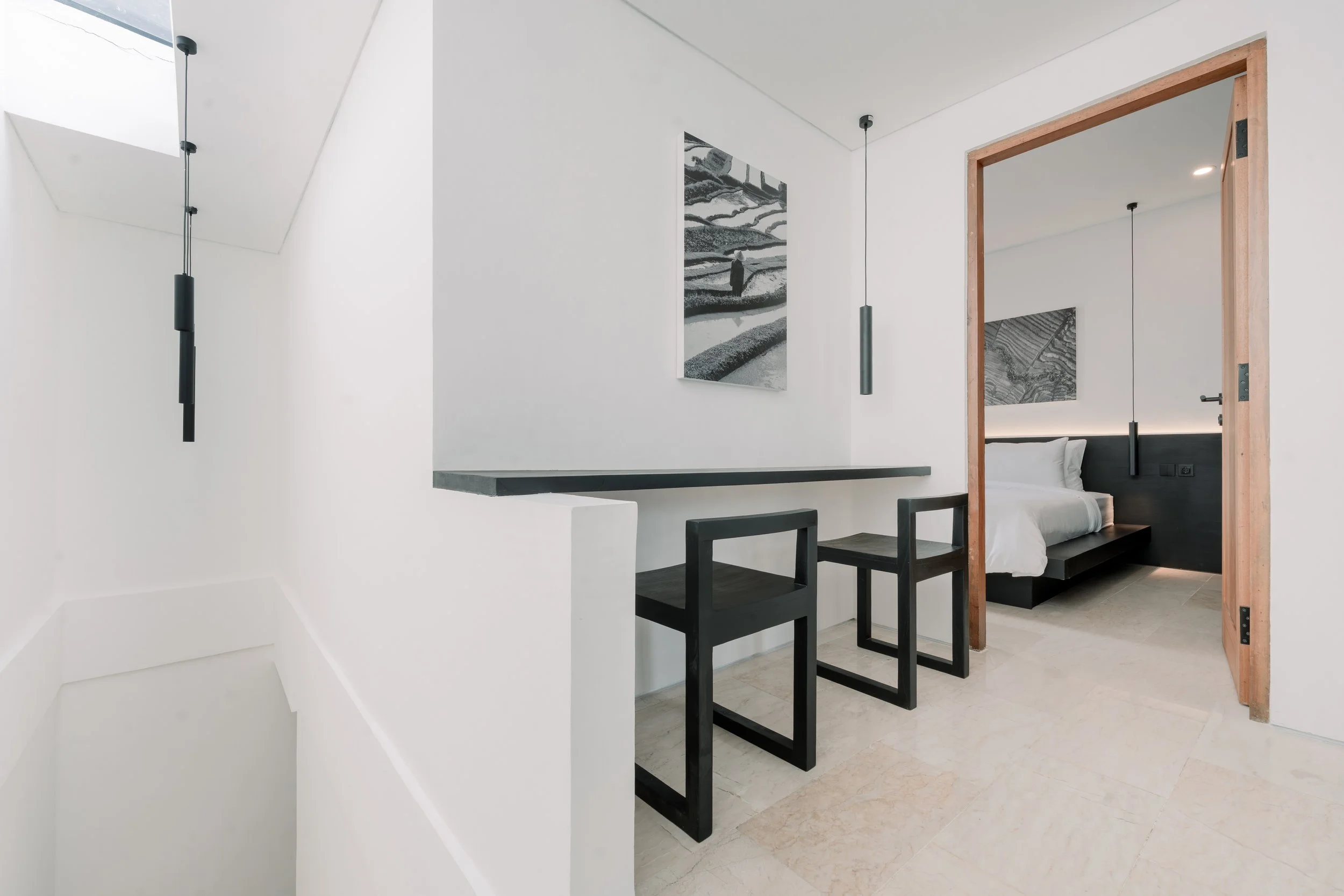 Minimalist hotel room with a sitting area, black chairs, and a view into the bedroom with white bedding and black accents.