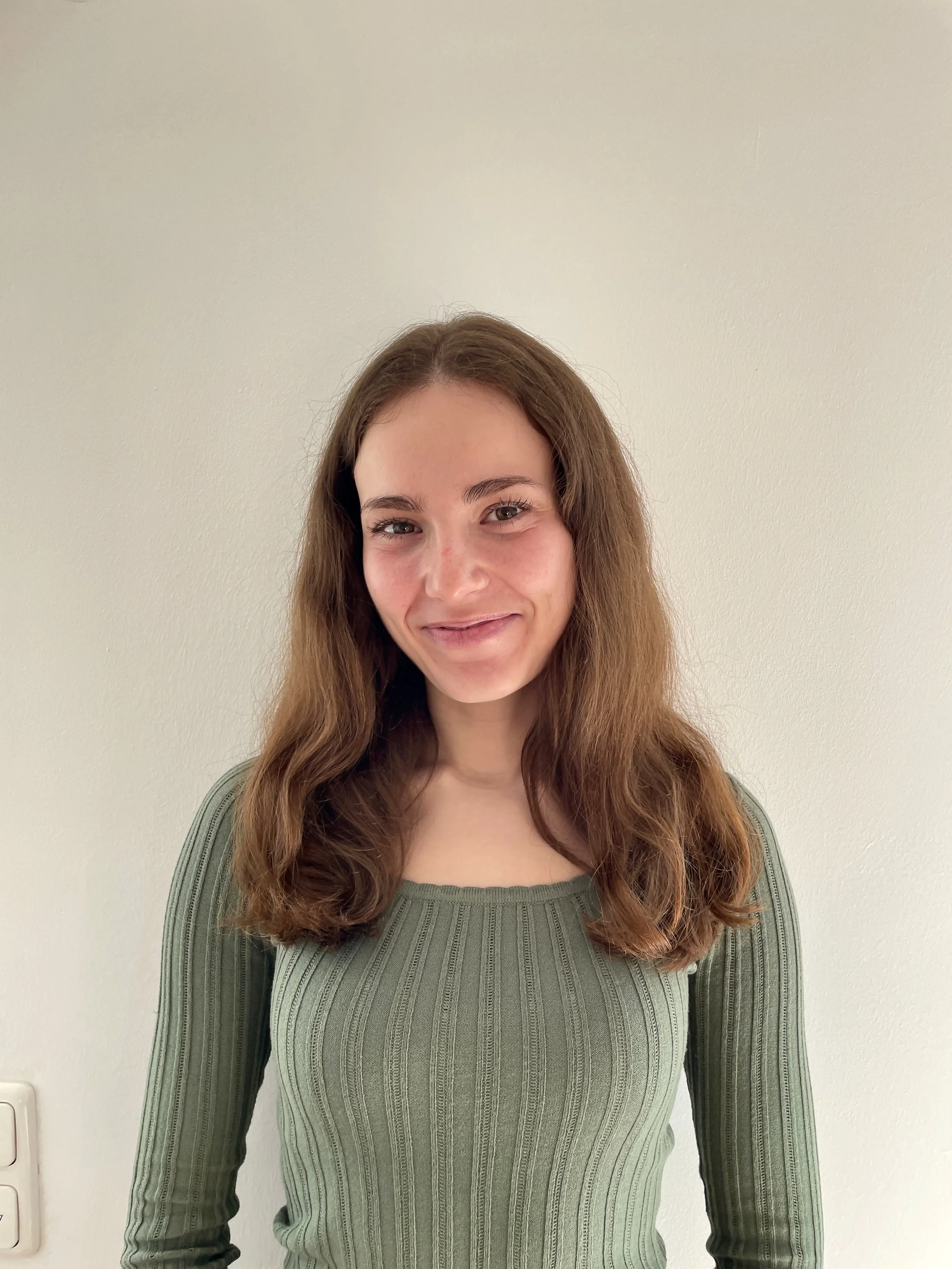 A young woman with long, wavy brown hair and light skin, smiling and wearing a long-sleeved green ribbed sweater. A motivated tutor.