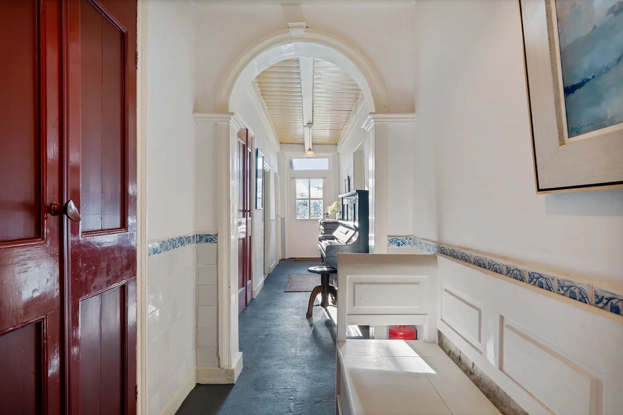 A narrow hallway with a white bench on the right, a red door on the left, and a window at the end of the hall letting in natural light. There is a piano near the window on the right and framed artwork on the walls.