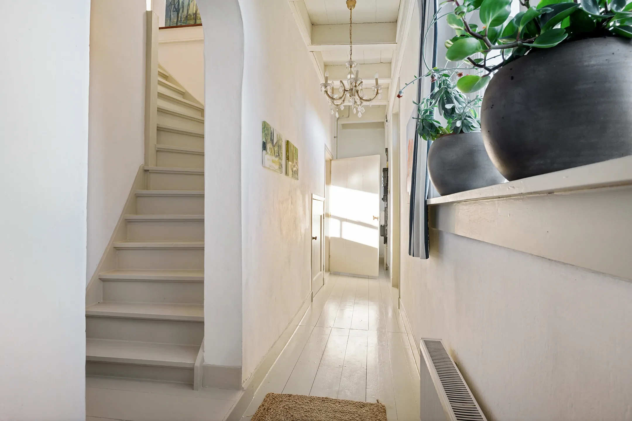 Bright hallway with white walls, wooden floor, a staircase on the left, potted plants on the right window sill, and a chandelier hanging from the ceiling. A door at the end of the hallway is open, allowing sunlight to stream in.