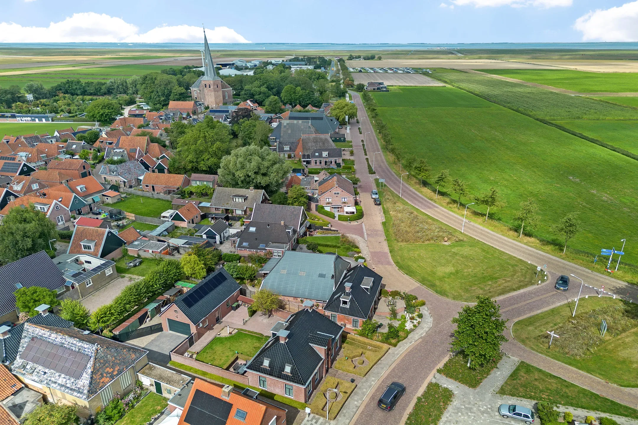 Aerial view of a small town with houses, a church with a tall steeple, green fields, and distant flat landscape under partly cloudy sky.