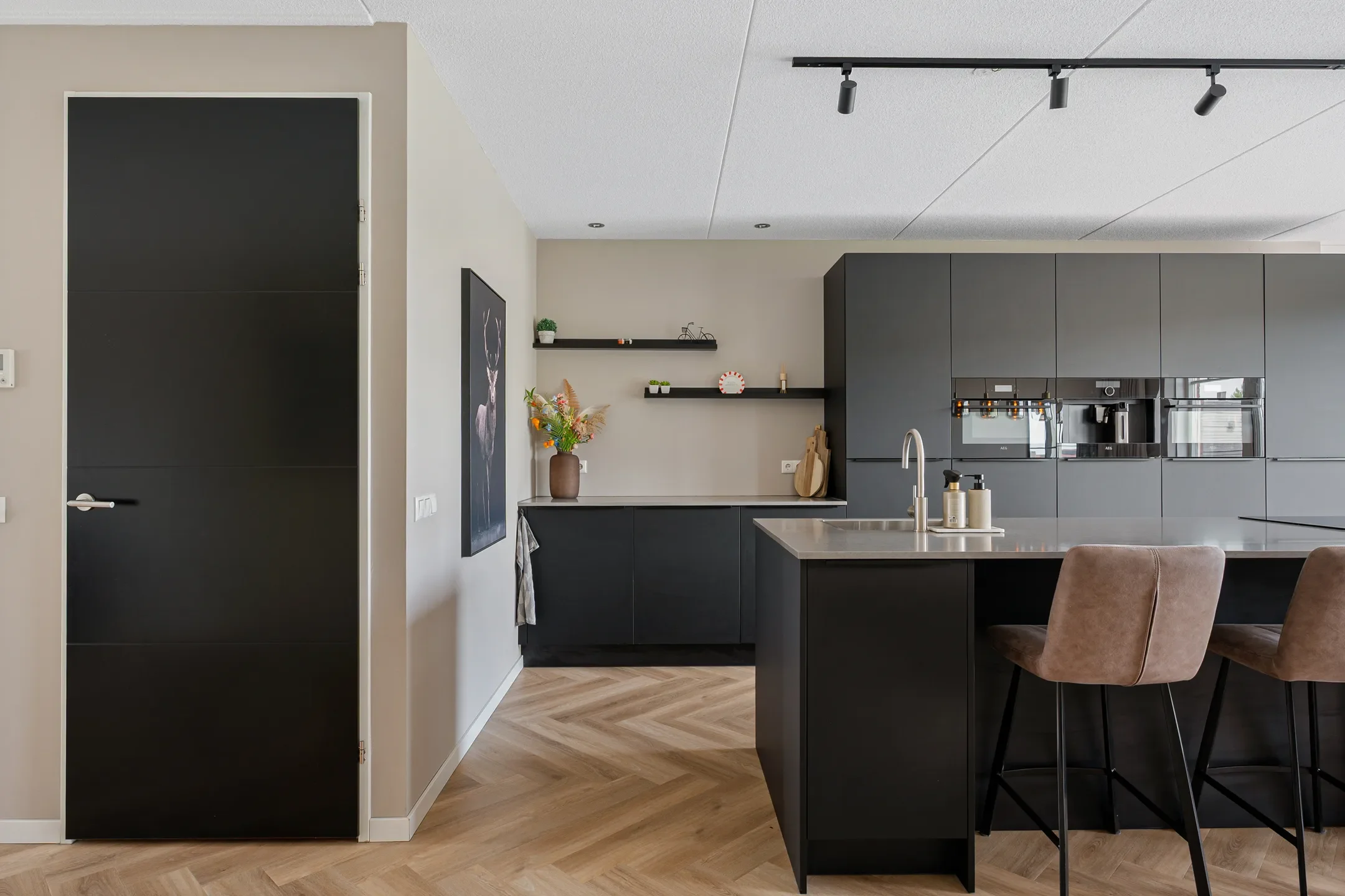 Modern kitchen with black cabinets, beige walls, and a kitchen island with brown upholstered chairs. Open shelving with decorative items and a vase with flowers.