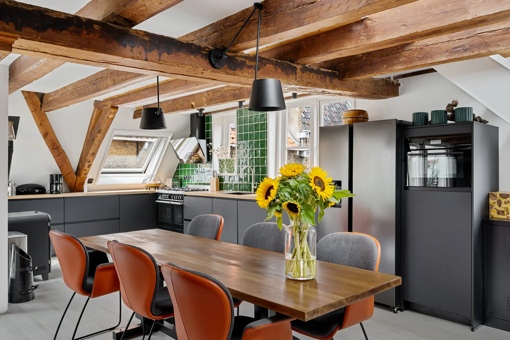 Modern kitchen and dining area with exposed wooden beams, black cabinets, a green tiled backsplash, and a wooden dining table with mixed orange and gray chairs. A vase of sunflowers is on the table.
