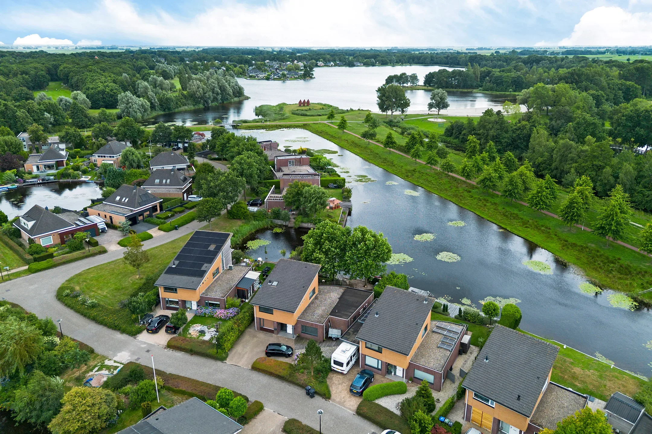 Aerial view of a residential neighborhood with modern houses, a river, and lush green trees, under a cloudy sky.