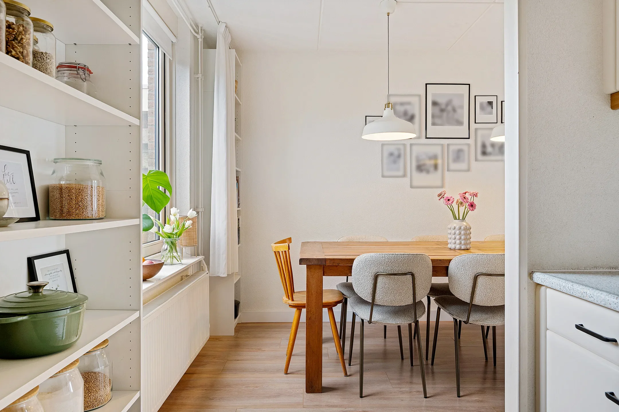 A cozy dining area with a wooden table, surrounded by a mix of four upholstered chairs and one wooden chair. The table has a white textured vase with pink flowers. A photo gallery decorates the white wall behind the table, and a white hanging pendant