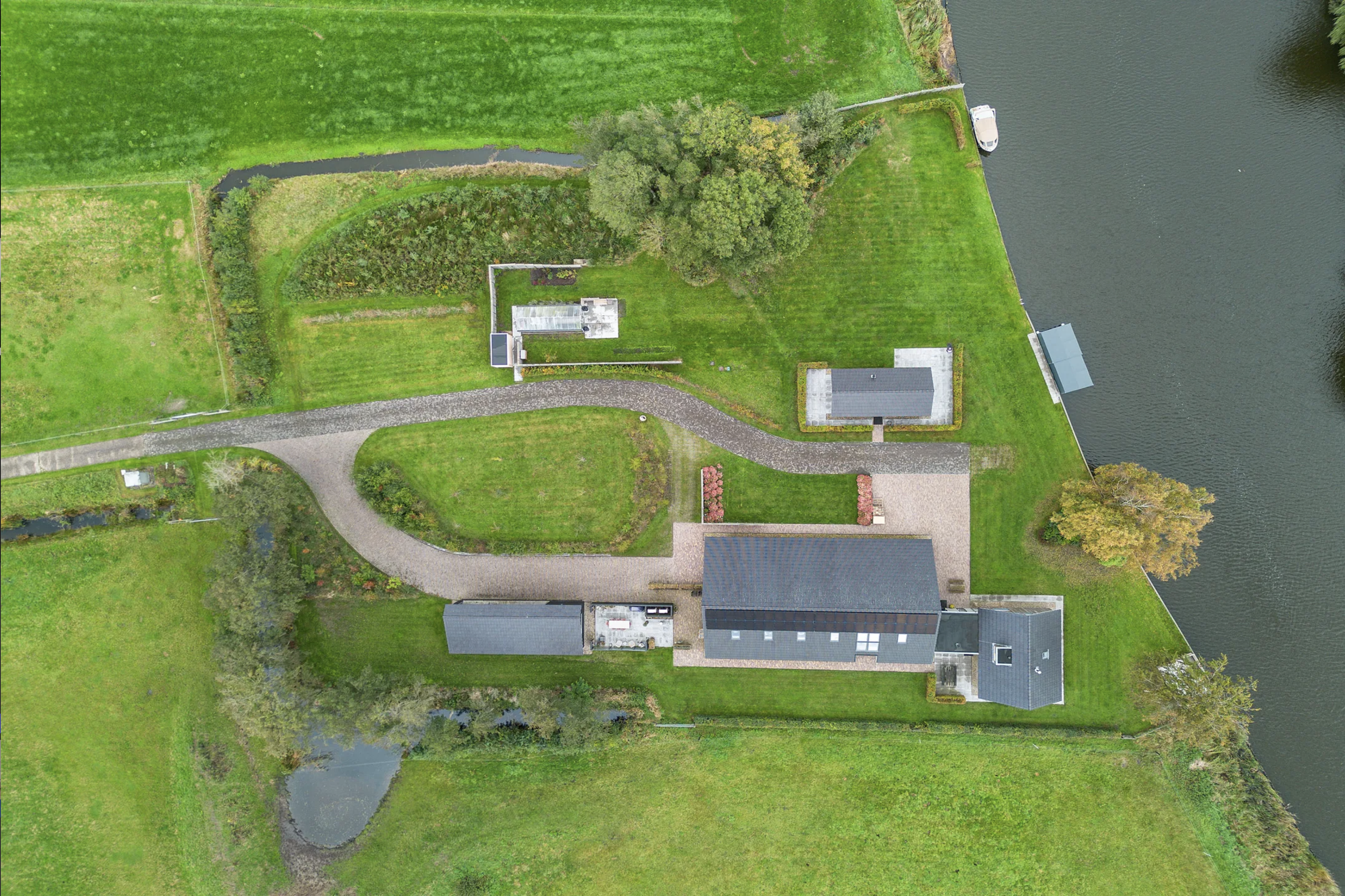 Aerial view of a large house with a dark roof, a gravel driveway, green lawn, and a pond with a dock on a waterway, surrounded by trees and open green fields.