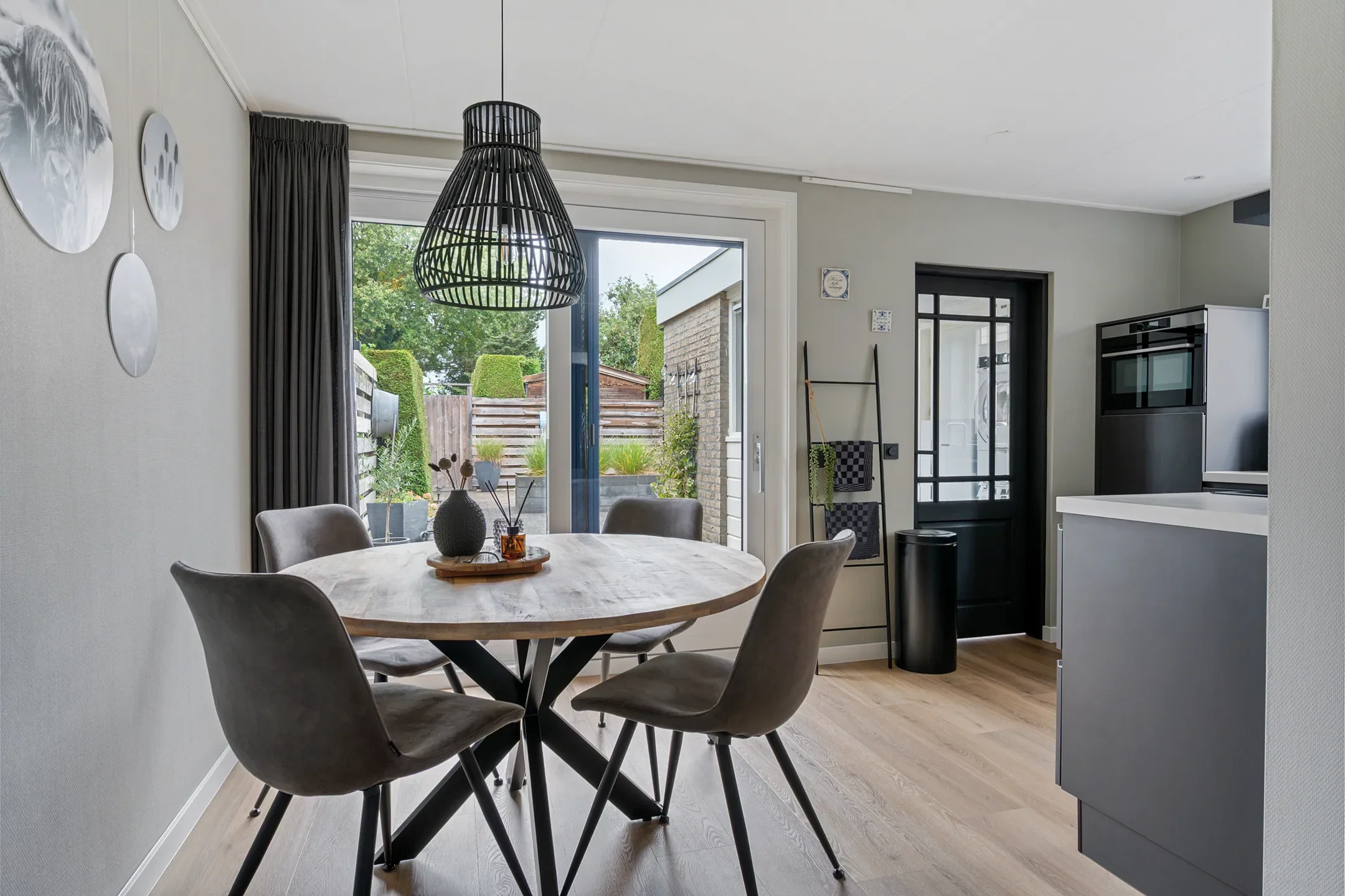 Dining area with a round wooden table and four gray chairs, large sliding glass door leading to outdoor patio, black hanging pendant light, minimalist decor with wall art, and view of a backyard garden.