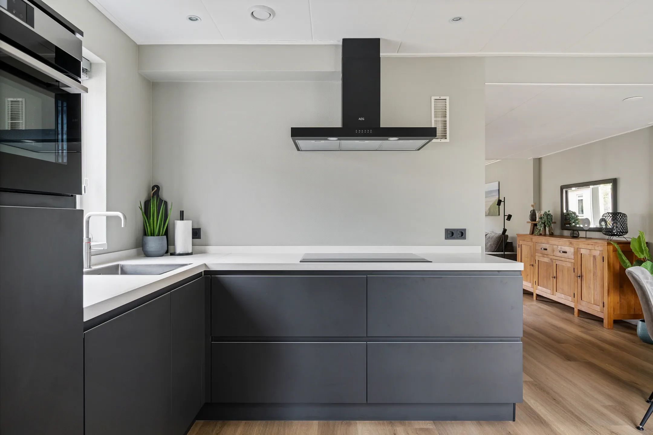 Modern kitchen with dark lower cabinets, white countertops, a built-in stove, a black range hood, a small plant near the sink, and a living space visible in the background.