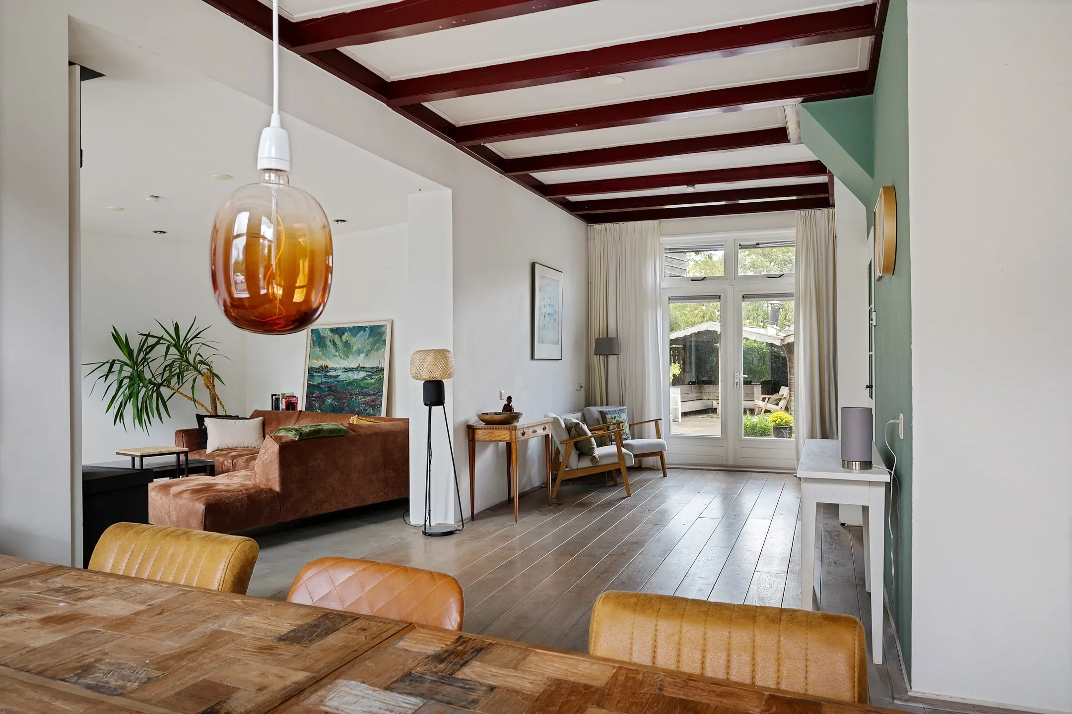 Living room with a view to an outdoor patio, featuring a brown sofa, large window with white curtains, and various chairs and lamps, with a wooden floor and a mid-century modern aesthetic.