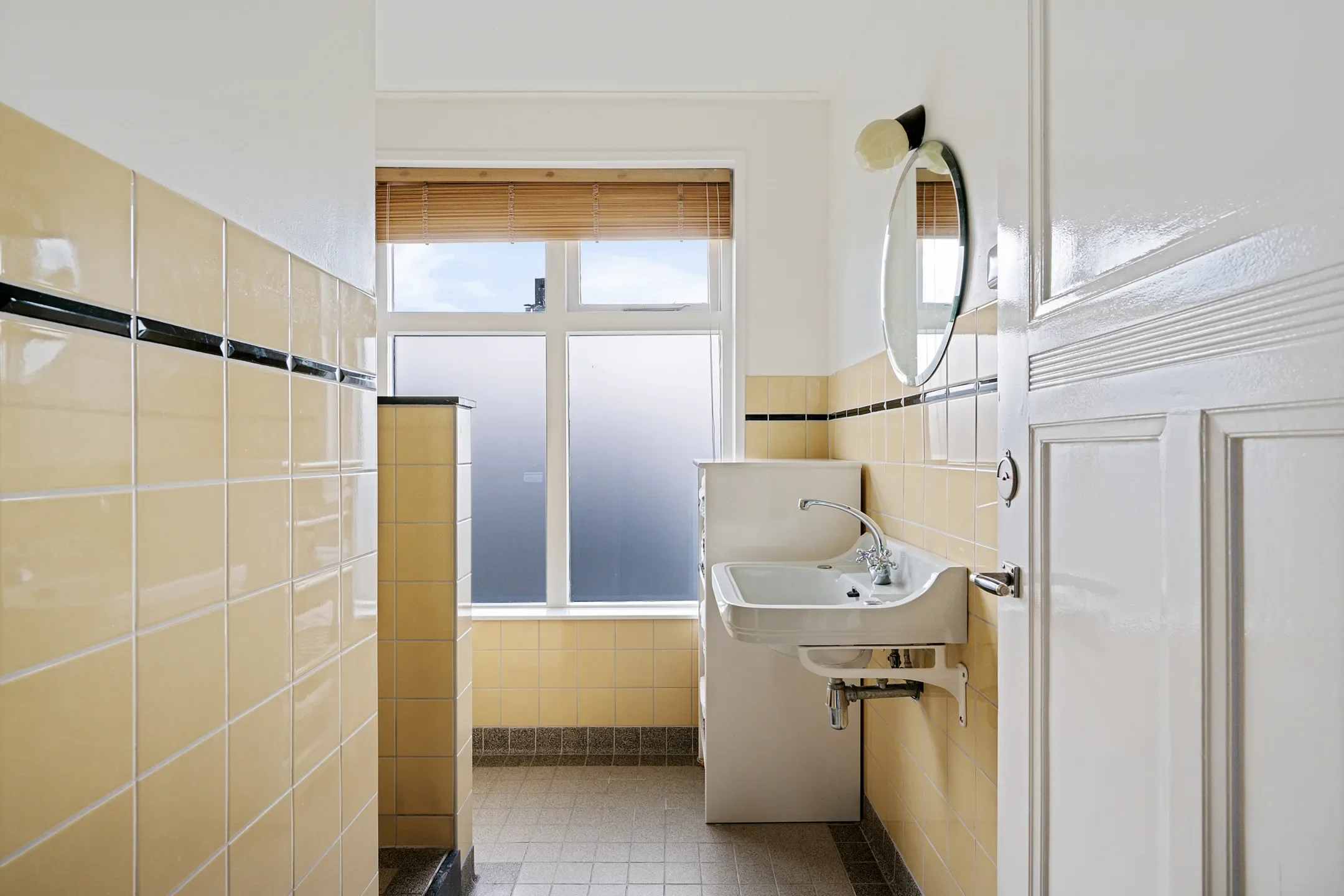 A small bathroom with yellow tiled walls, a white sink with a shelf, a small mirror above the sink, a frosted window with blinds, and a wooden door.