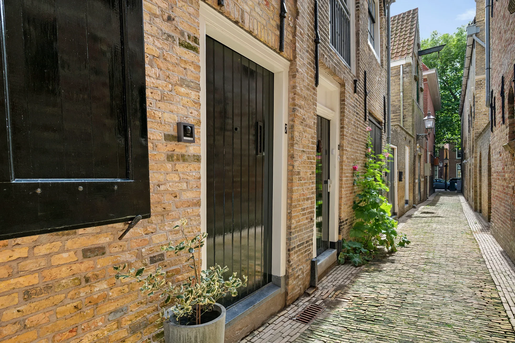 Narrow cobblestone alleyway with brick buildings, plants, and black-windowed doors.