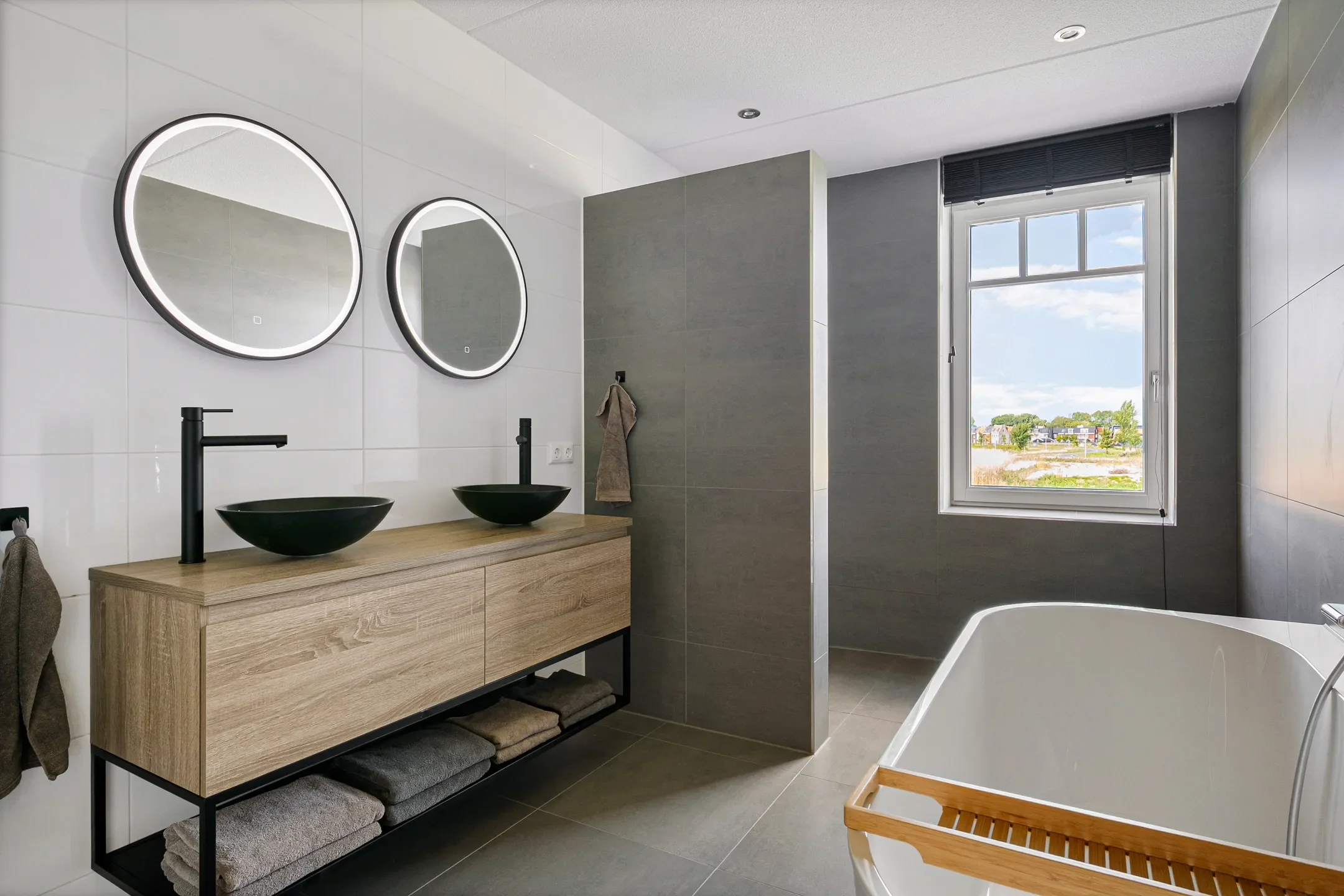 Modern bathroom with two black vessel sinks on a wooden vanity, two round mirrors with built-in lighting, a large window showing an outdoor landscape, and a bathtub with a wooden tray.