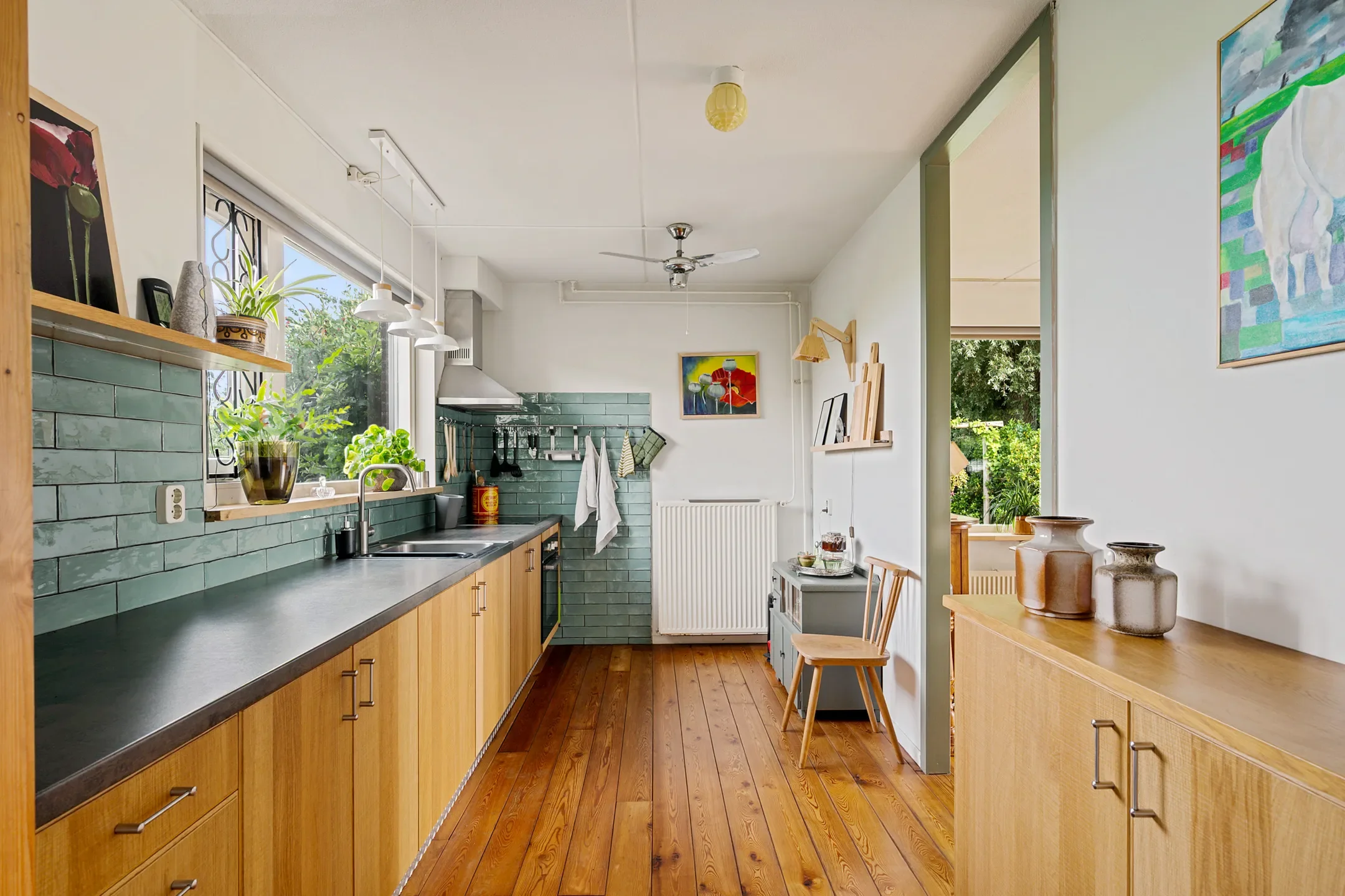 Kitchen with wooden cabinets, green tiled backsplash, and a large window with plants outside. There are hanging lights, a sink, and small decor items on the shelves.