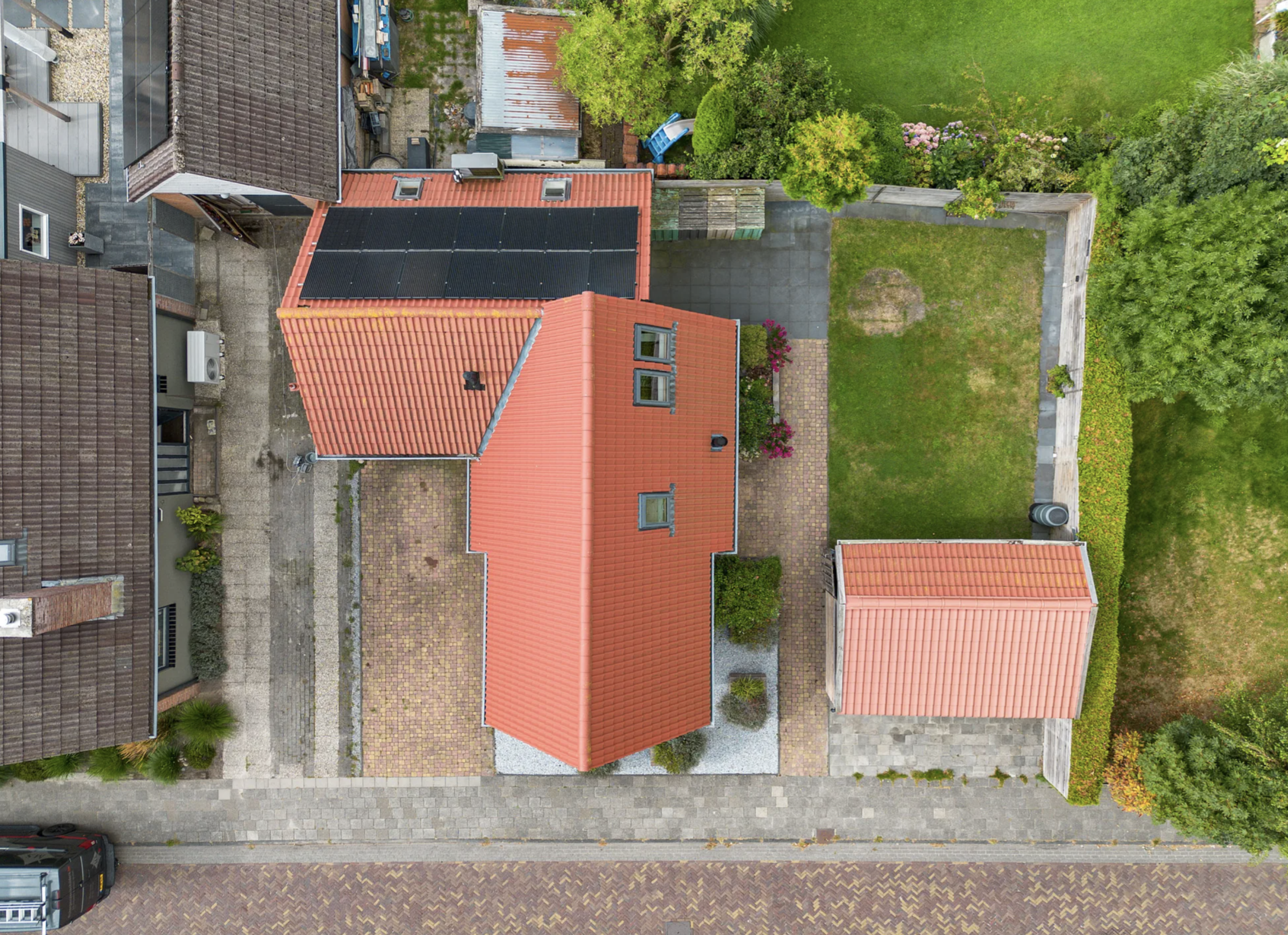 Aerial view of a suburban backyard with a house, small outbuilding, paved pathways, a grass lawn, trees, and bordering walkway.