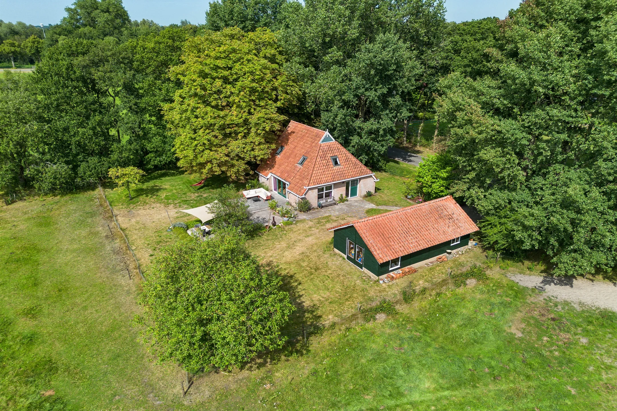 Aerial view of a house and a separate garage/building surrounded by green trees and grassy yard, with outdoor furniture and a canopy in the yard.