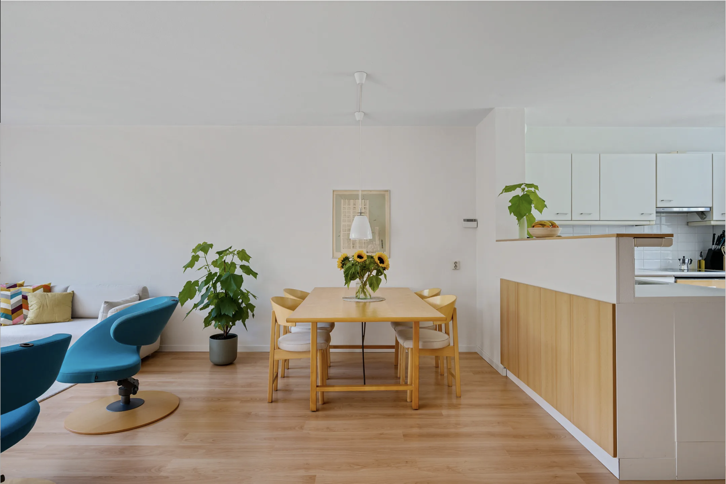 Open-concept living and dining area with a wooden dining table, six chairs, and a bouquet of sunflowers. The space includes a kitchen with white cabinets and a counter with a potted plant. There are two turquoise chairs on the left and a light-colore