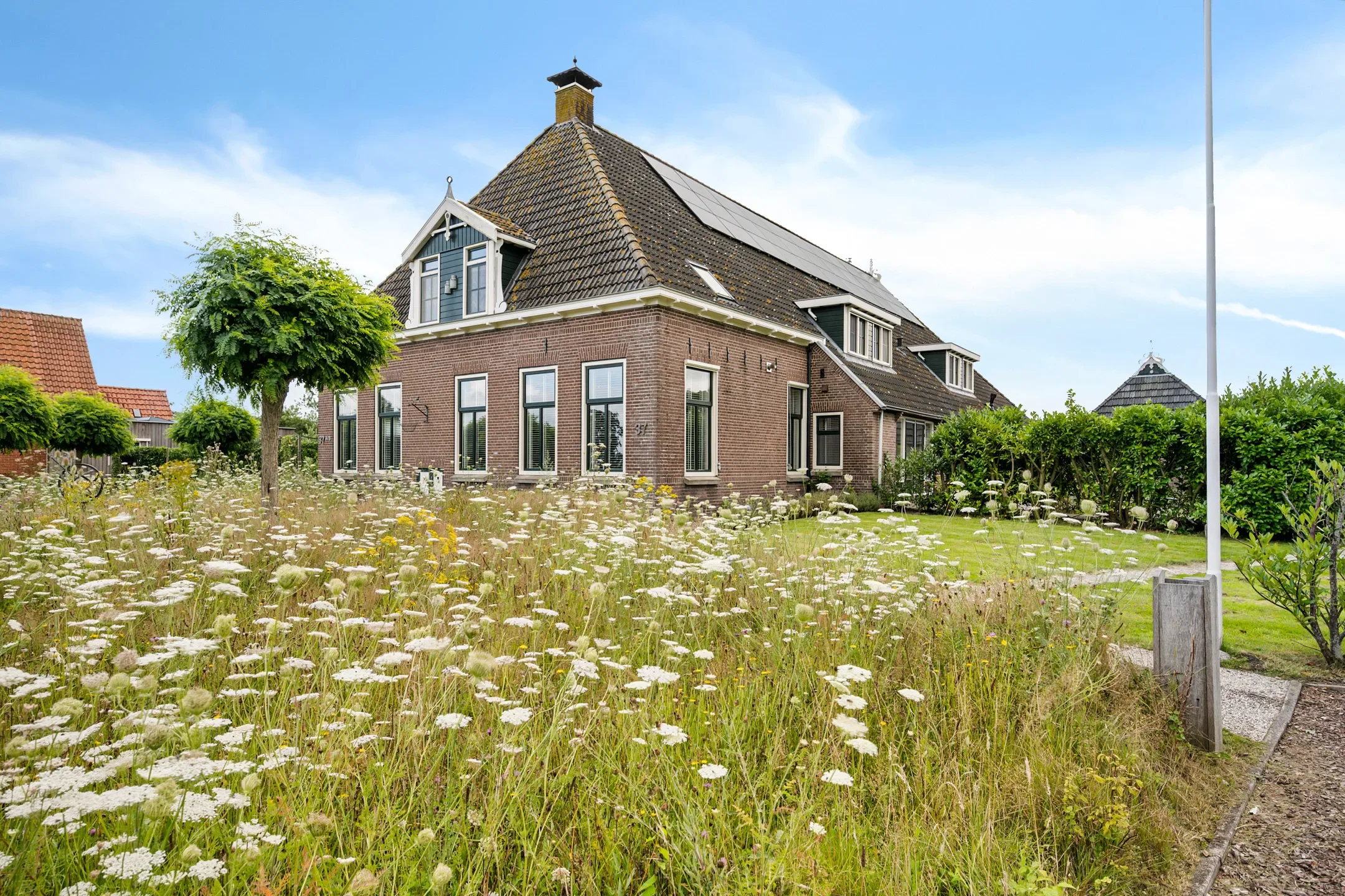 A large brick house with a steep shingled roof, solar panels, and multiple windows, surrounded by a lush garden with tall grass, wildflowers, and trimmed trees under a partly cloudy sky.