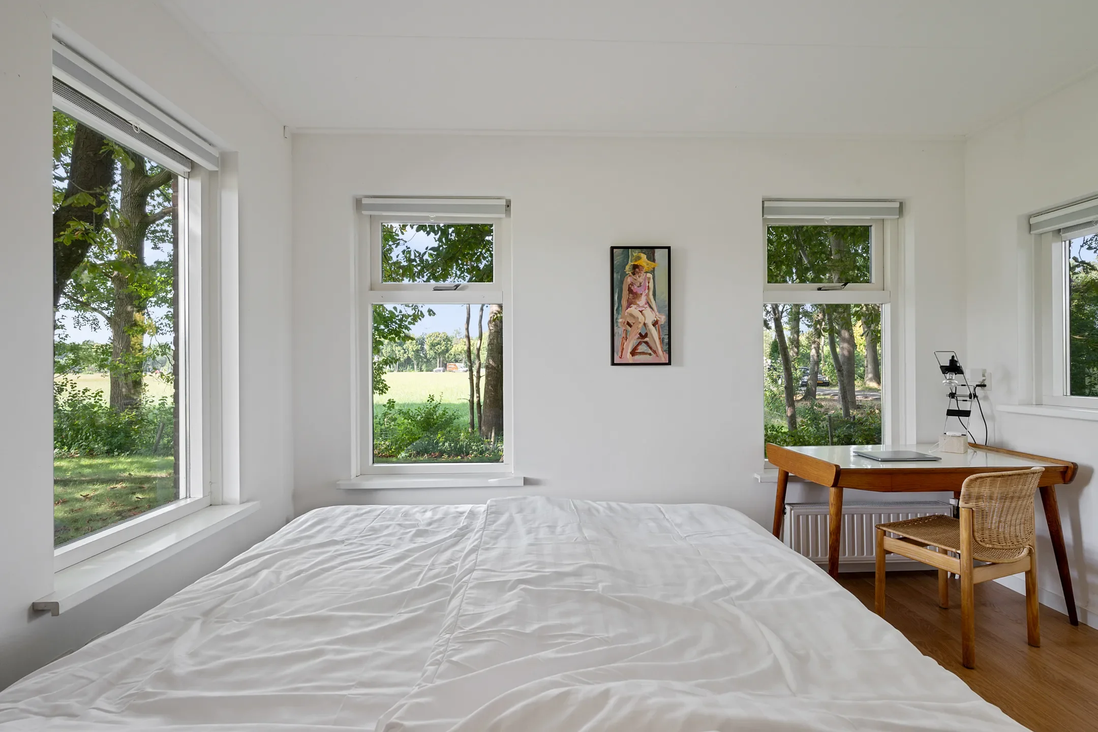 A bright bedroom with white walls and large windows showing green trees outside. A bed with white bedding, a wooden desk with a wicker chair, a small telescope, and a framed artwork of a woman in a yellow hat are visible.