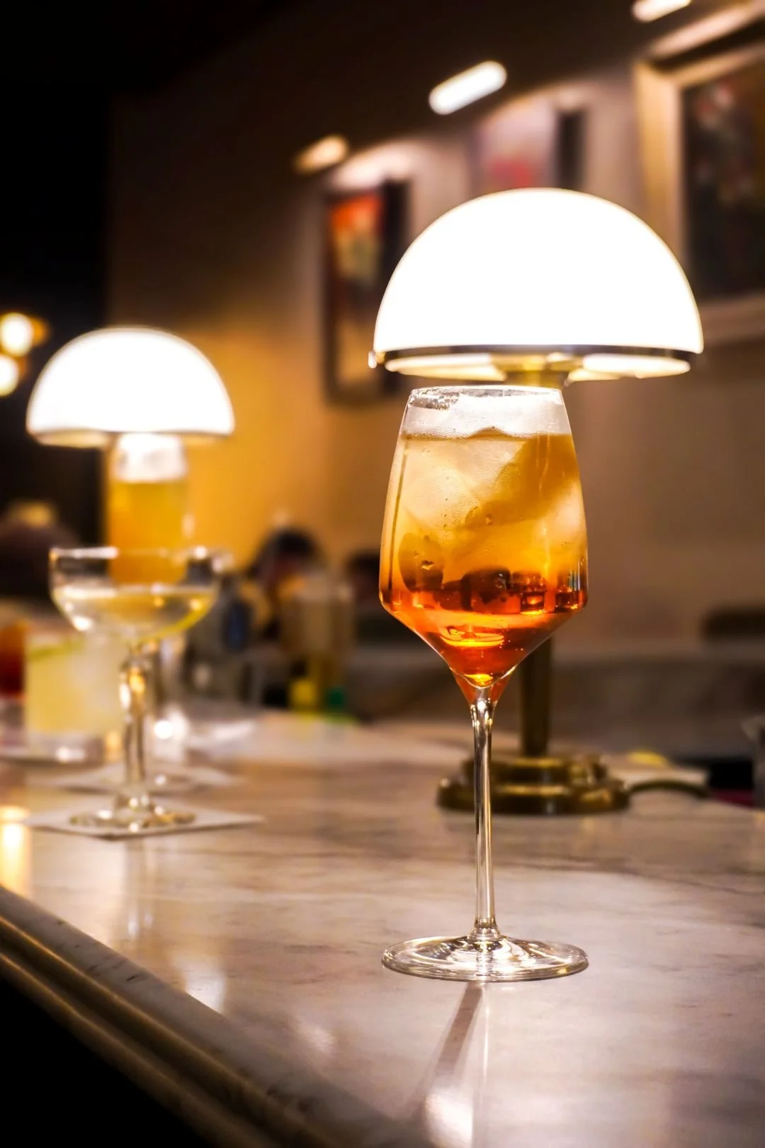 A cocktail in a tall glass with ice cubes and a reddish hue, placed on a bar counter with soft lighting and lamps in the background in Zinis pizza bar in Amersham, Bucks