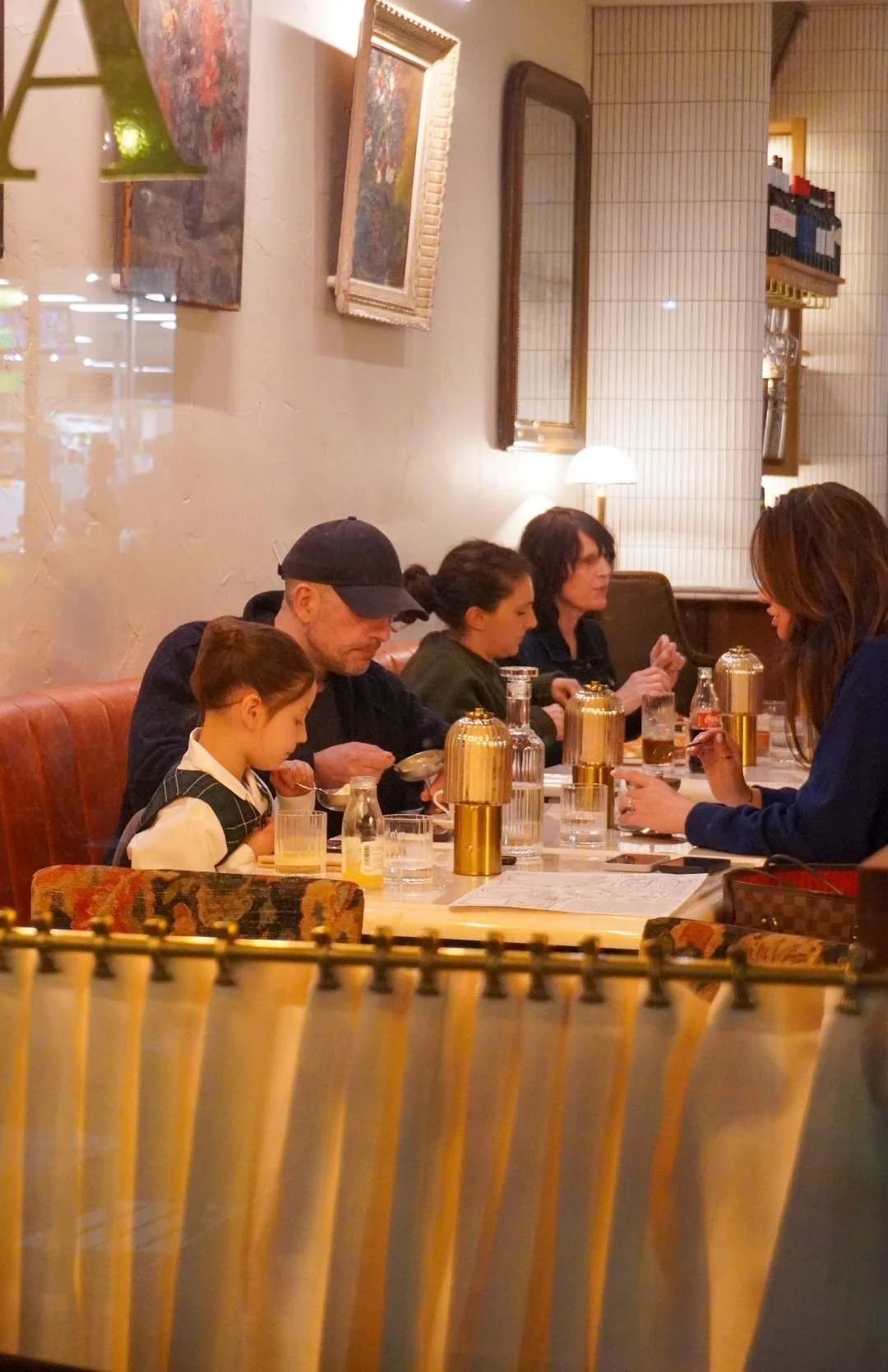 Family dining at a Zinis pizza bar in Amersham, Bucks, with a young girl, a man with a cap, two women, and bottles of water and drinks on the table, warm indoor lighting, decorated walls with paintings, mirror, and shelves.
