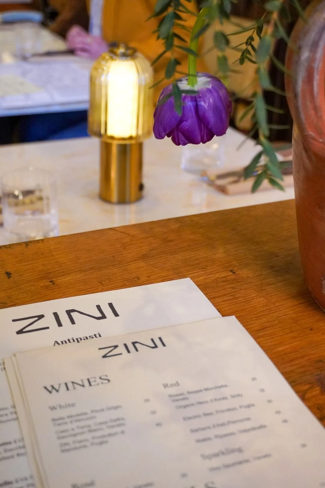 A restaurant table in Zinis pizza bar in Amersham, Bucks with a menu labeled "ZINI" and a purple flower in a vase, with a blurred background including a glass of water and a gold-colored lamp.