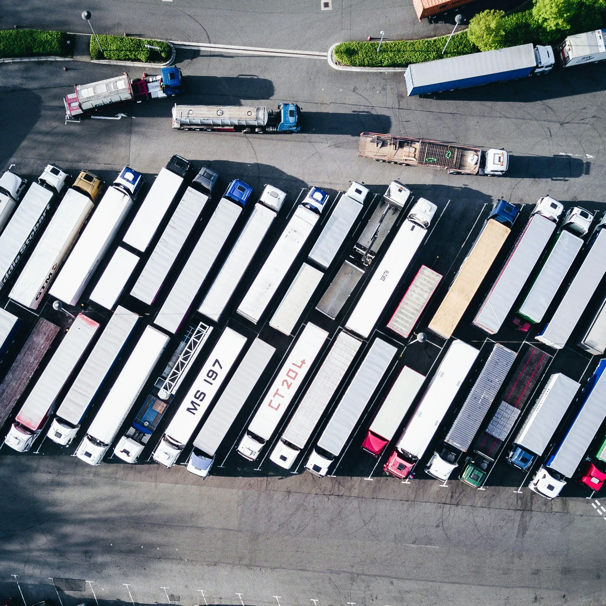 An aerial view of a parking lot filled with various trucks parked in rows. Some trucks are large with white or colored trailers, and there are other vehicles parked along the edge of the lot. The area is surrounded by pavement and some greenery.