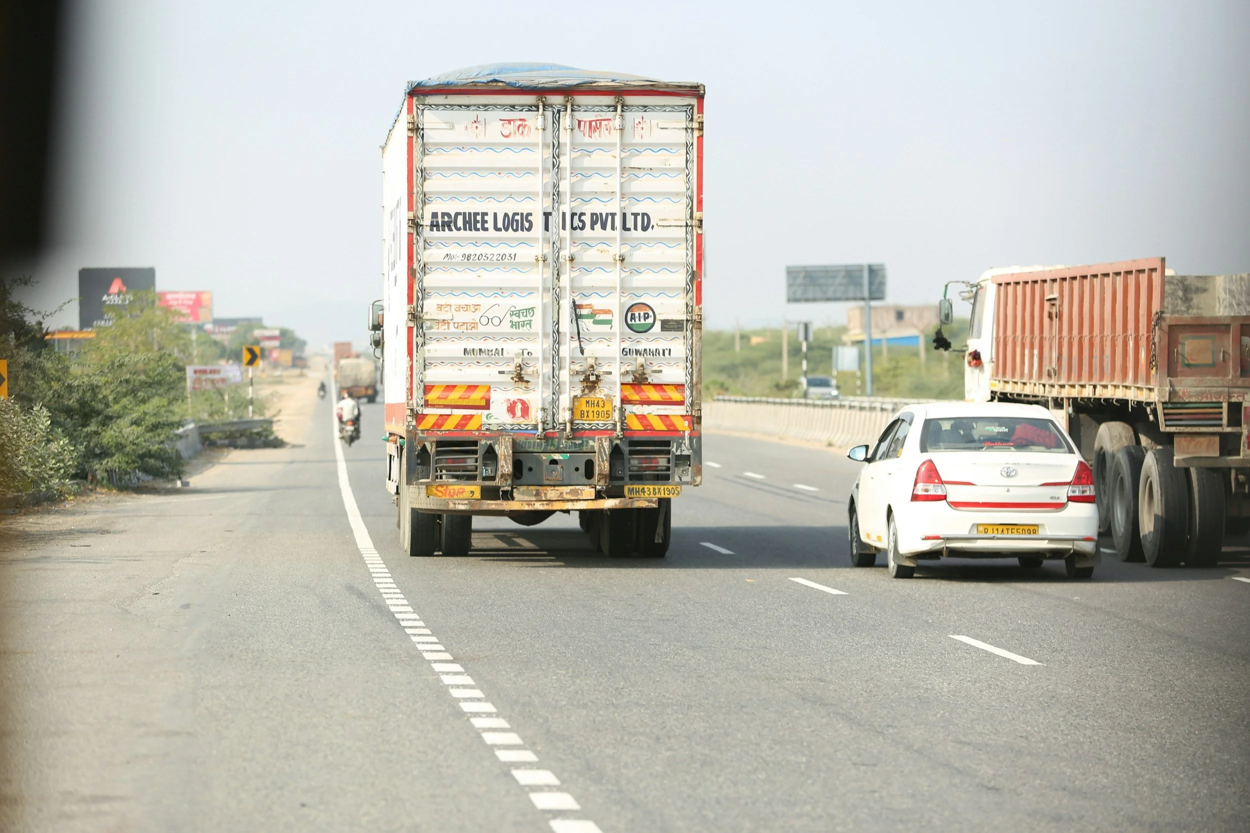 Vehicles traveling on a multi-lane highway, including a large truck, a white car, and a toyota truck, with roadside greenery and distant billboards under a cloudy sky.