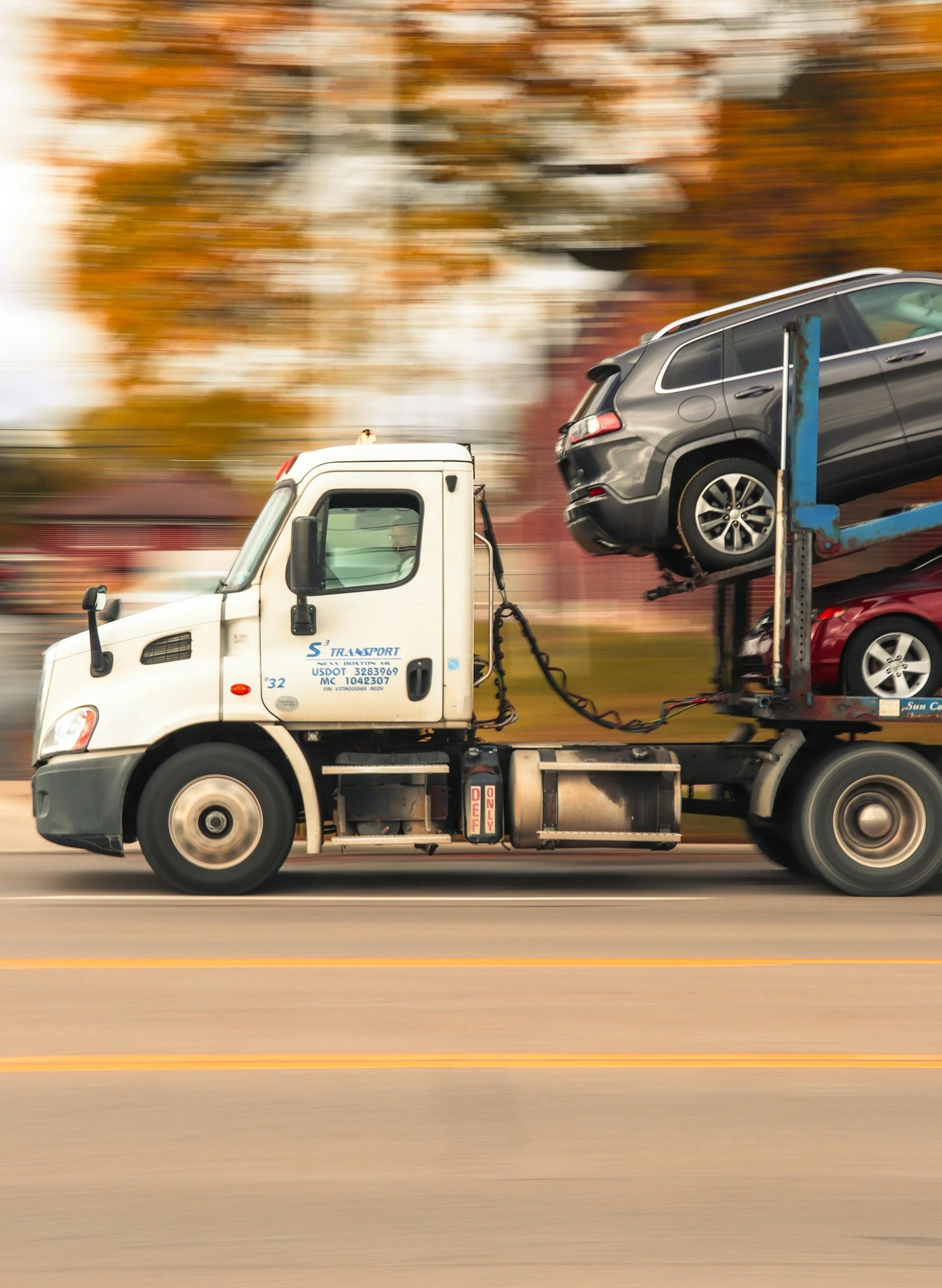 Tow truck carrying two parked cars, one black SUV on top and one red car underneath, driving down a road with autumn trees in the background.