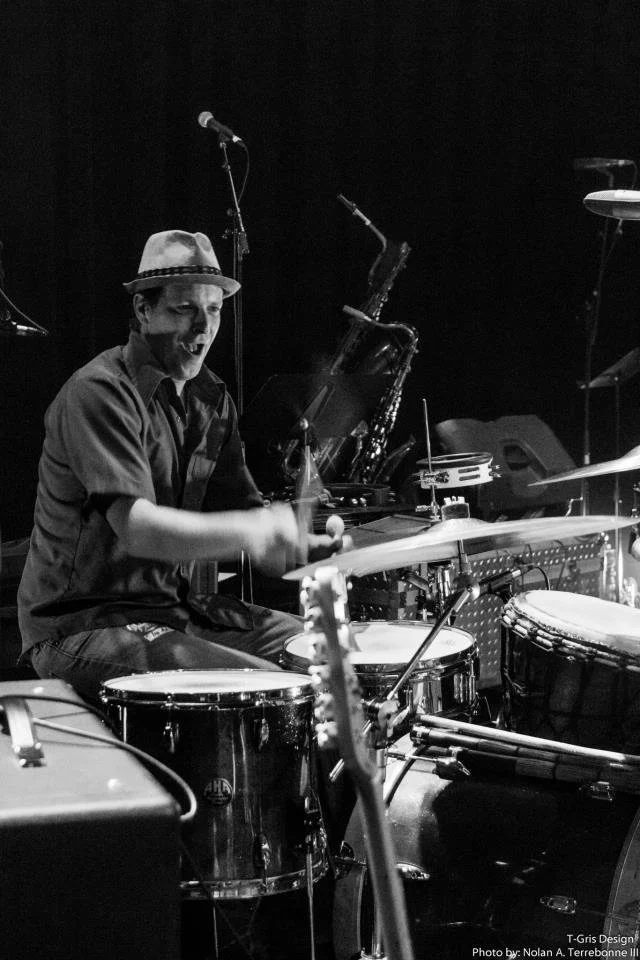 A drummer playing percussion instruments on a stage with a microphone and saxophone nearby in a black and white photo.