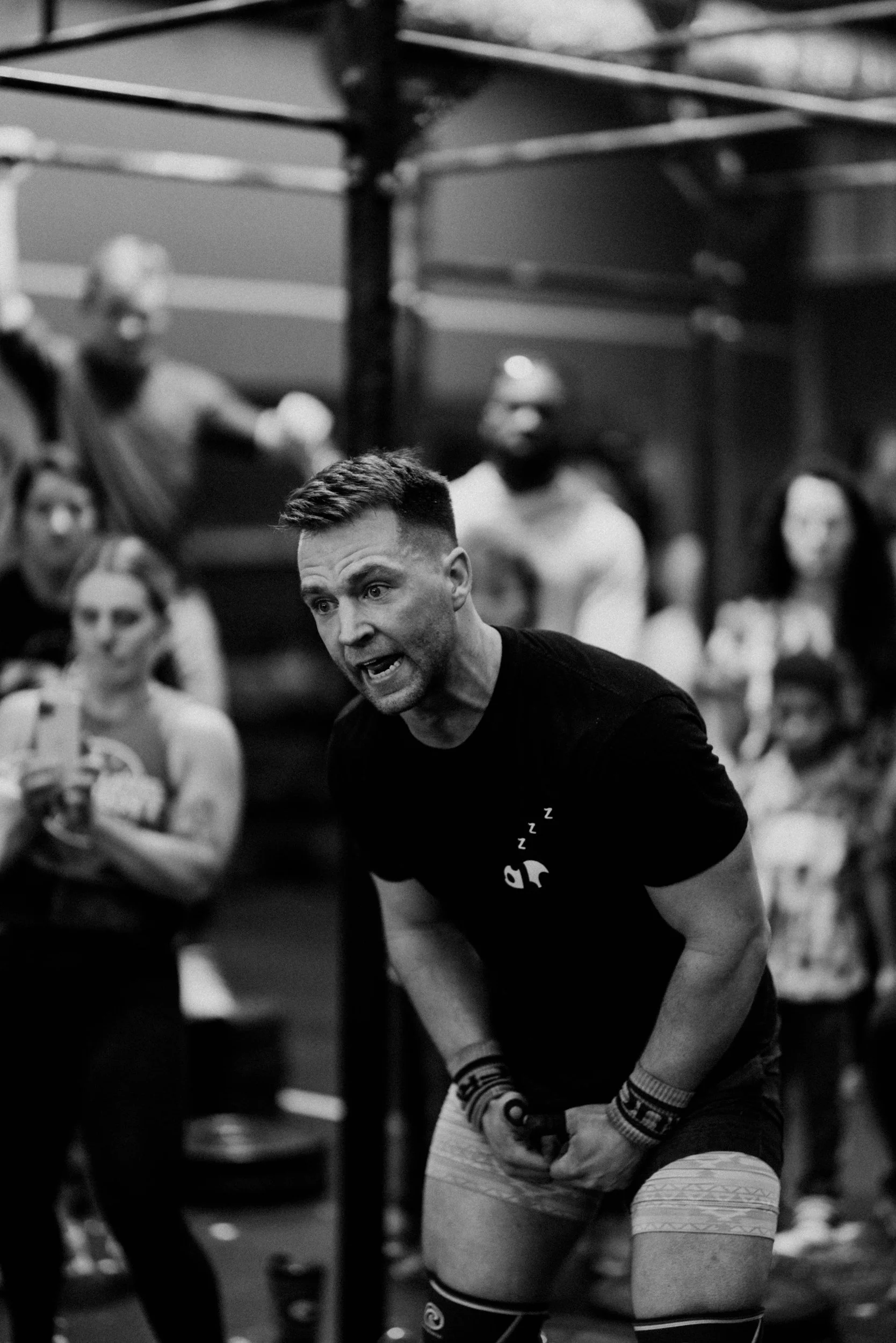 A man appears to be shouting or coaching during a CrossFit workout, surrounded by a group of people in a gym.