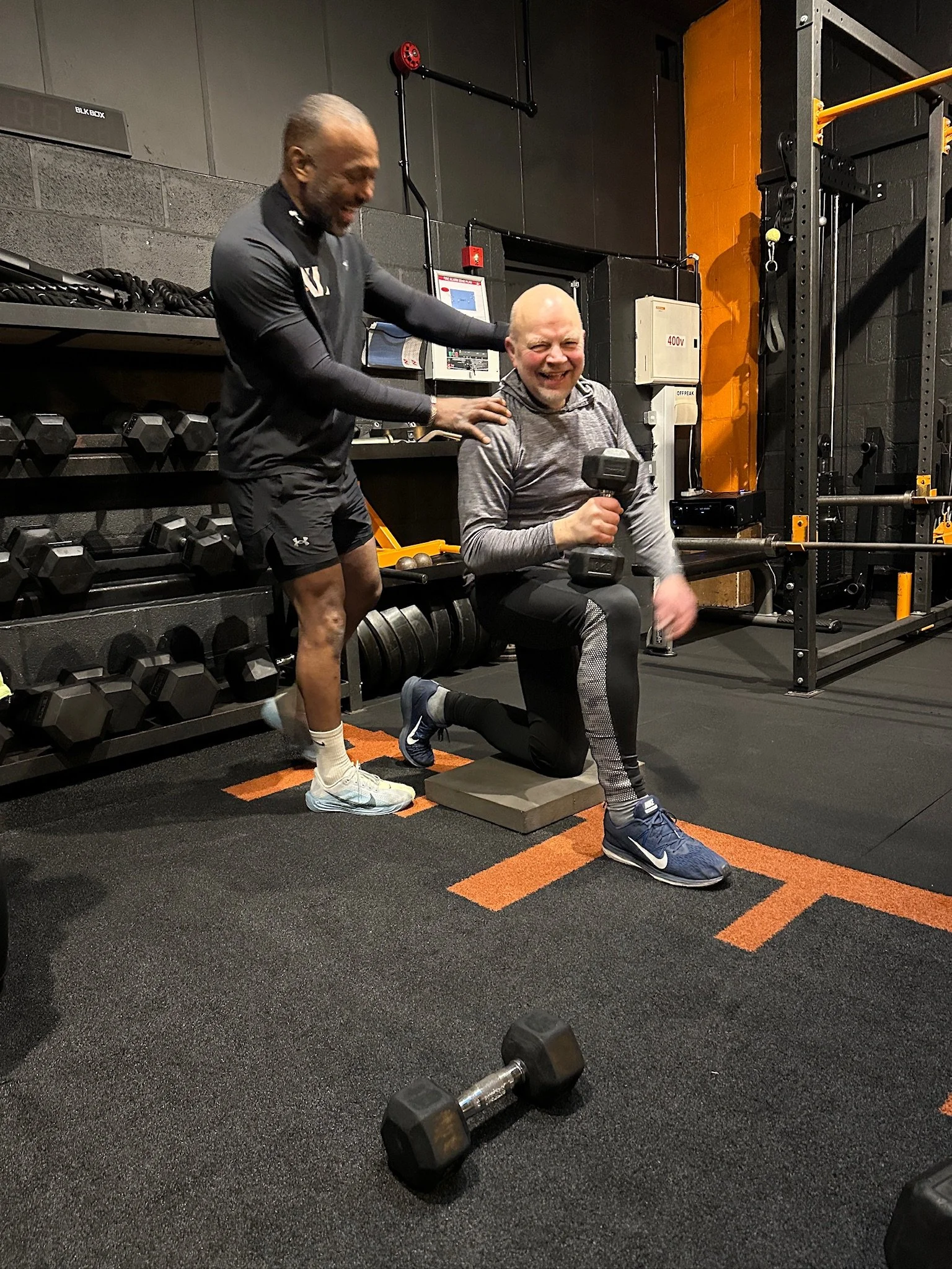 Two men at a gym; one kneeling on a cushion holding a dumbbell, smiling, while the other man standing beside him, touching his shoulder and smiling.