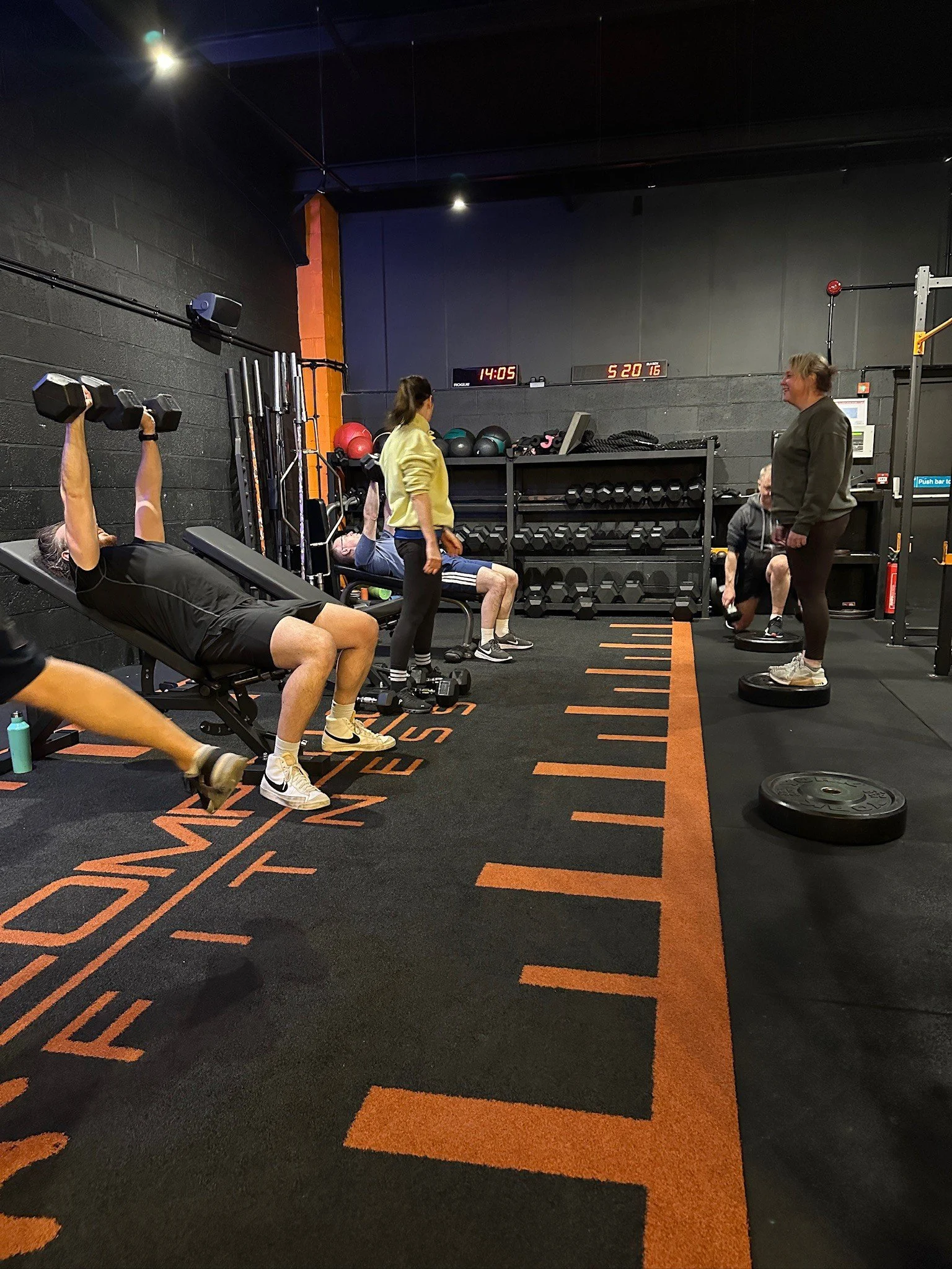 People working out at a gym with black walls, orange accents, and shelves of weights, with a coach standing on a balance trainer.