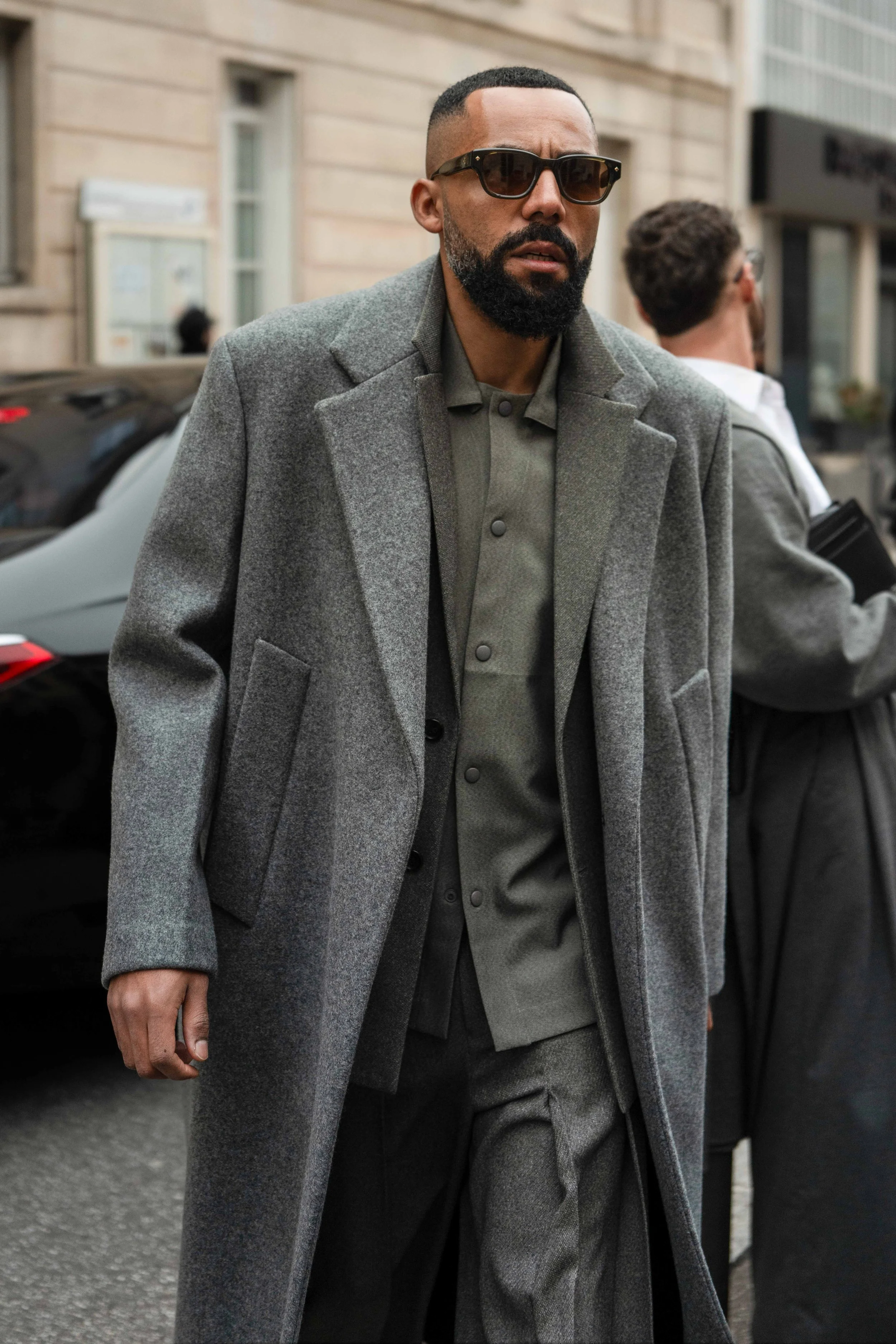 Street Style Photo of Jean-Claude Mpassy before the Issey Miyake show during Paris Fashion Week. He wears a layered look featuring a grey coat from Jil Sander and a shirt and blazer from Issey Miyake.