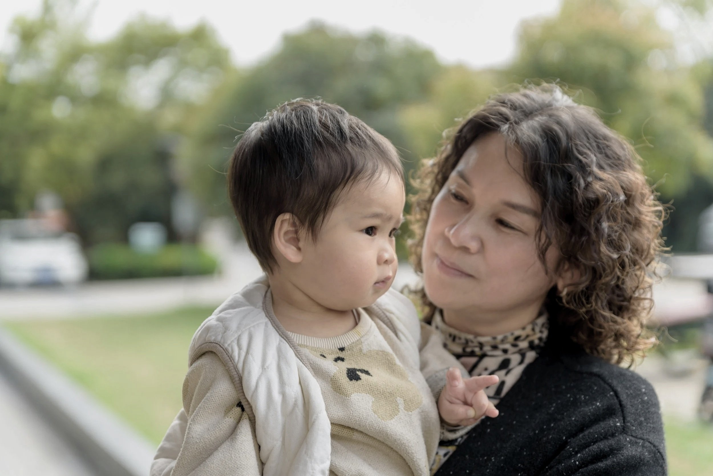 A woman with curly hair holding a young boy with dark hair outdoors in a park. They are close to each other, and the woman is looking at the boy while the boy looks to the side. Green trees and blurred cars are in the background.